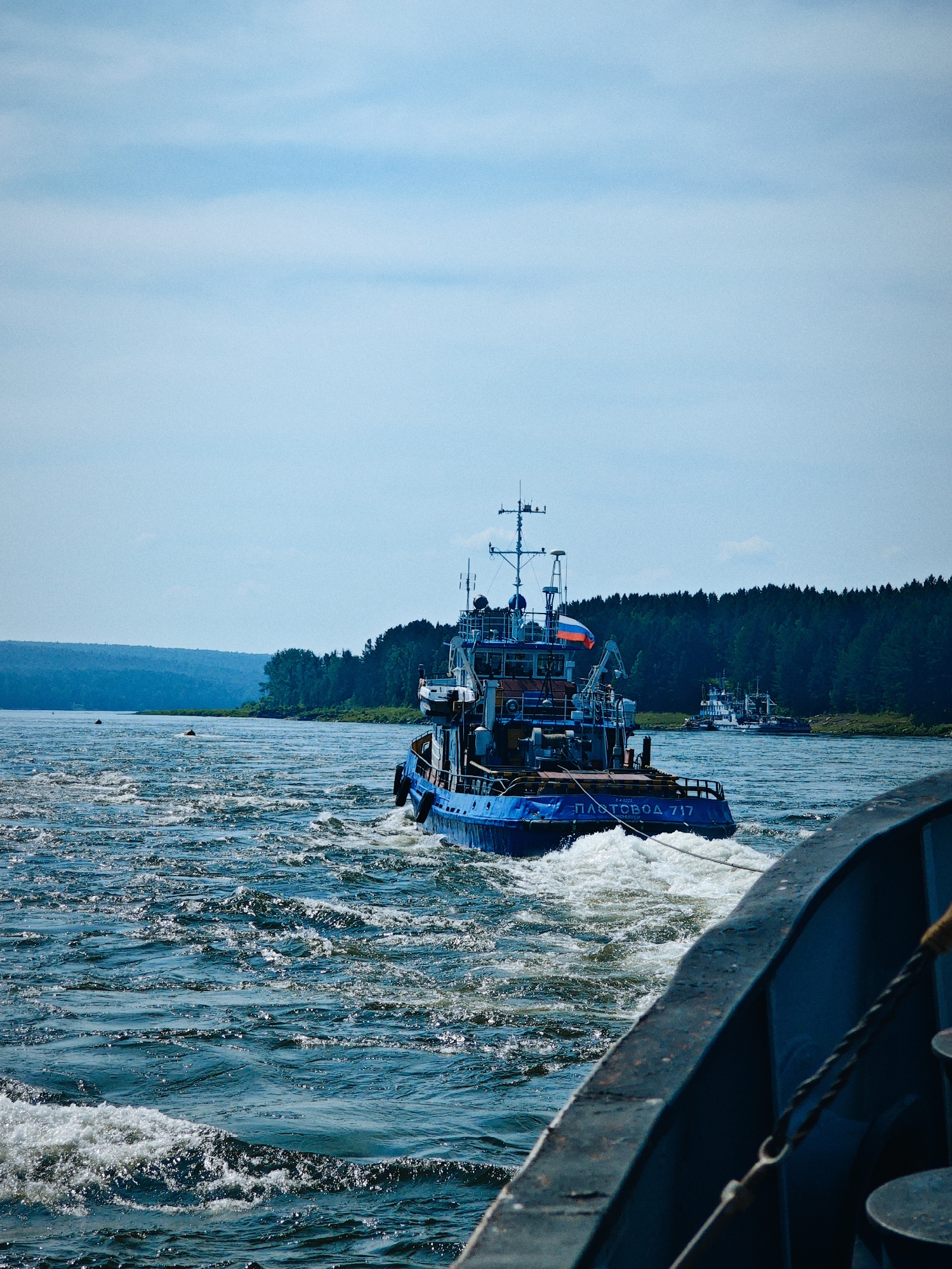 A blue tugboat, identified by the Russian text "ПЛАТОBOД 717" on its stern, is seen from the perspective of another vessel in the foreground. The tugboat is creating a significant wake as it moves through choppy, dark blue water. In the distance, a dense, dark green forest lines the far bank of a wide river, meeting a hazy, blue sky. Two buoys are visible in the water between the two vessels. Another, smaller white vessel is further in the distance, behind the tugboat. The overall scene suggests a sunny day on a Russian river.