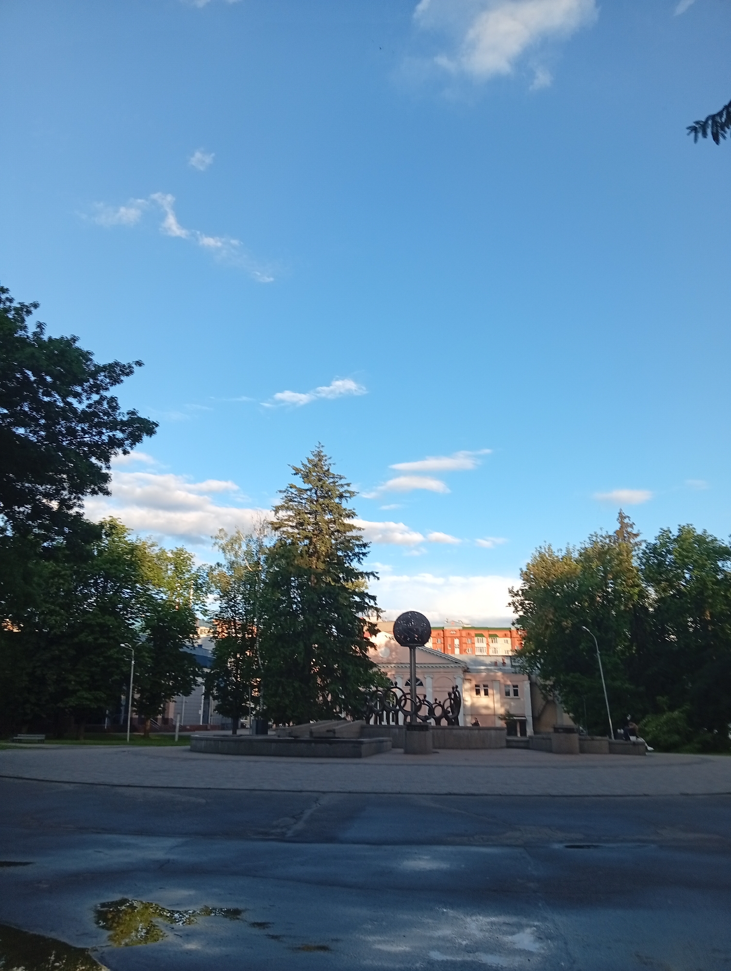 The image shows a street scene in Poltava, Ukraine. In the foreground, there is a wet, asphalt street reflecting the sky and trees. Behind this is a paved square with a low stone structure. On the structure is a sculptural composition, possibly a monument or art installation. The composition features a sphere on a pole, surrounded by silhouetted figures connected by a circular design. In the background, there are trees and buildings, including a pink building with white trim and windows. The sky above is bright blue with scattered white clouds.