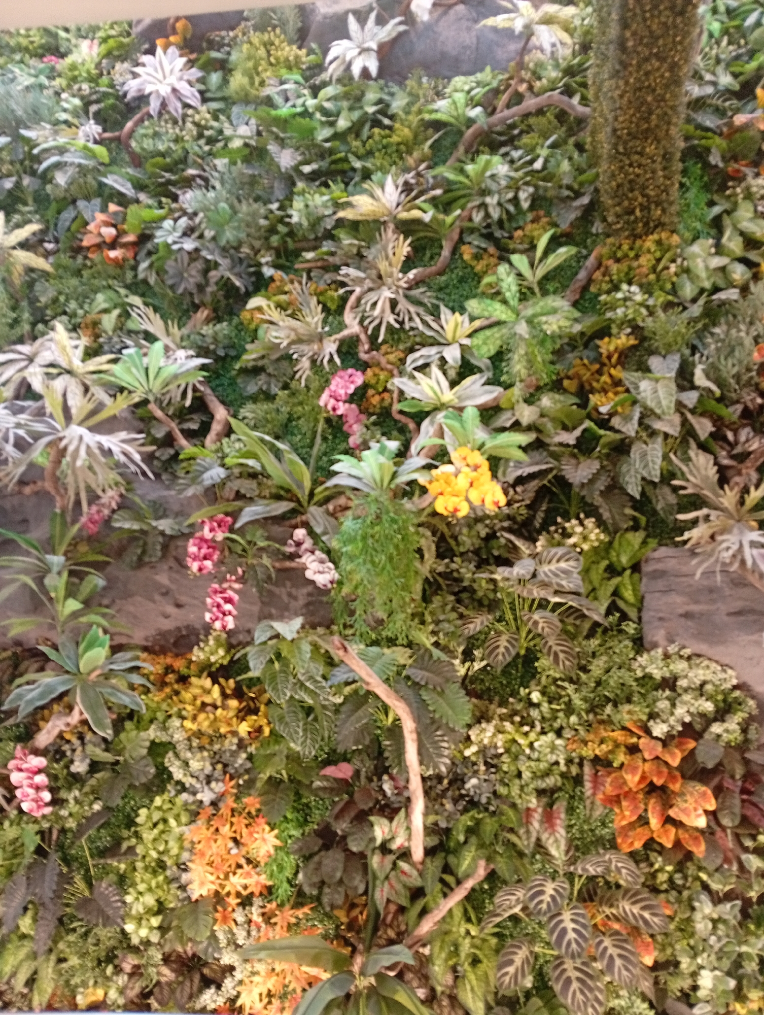 The image captures a lush vertical garden wall located in Denpasar, Indonesia. The wall is densely covered with a diverse range of plant species exhibiting various textures, colors, and leaf shapes. Among the foliage, there are white, yellow, pink, and orange flowers, adding pops of color. Wooden branches are interwoven among the plants. The backdrop behind the wall reveals grey stone.