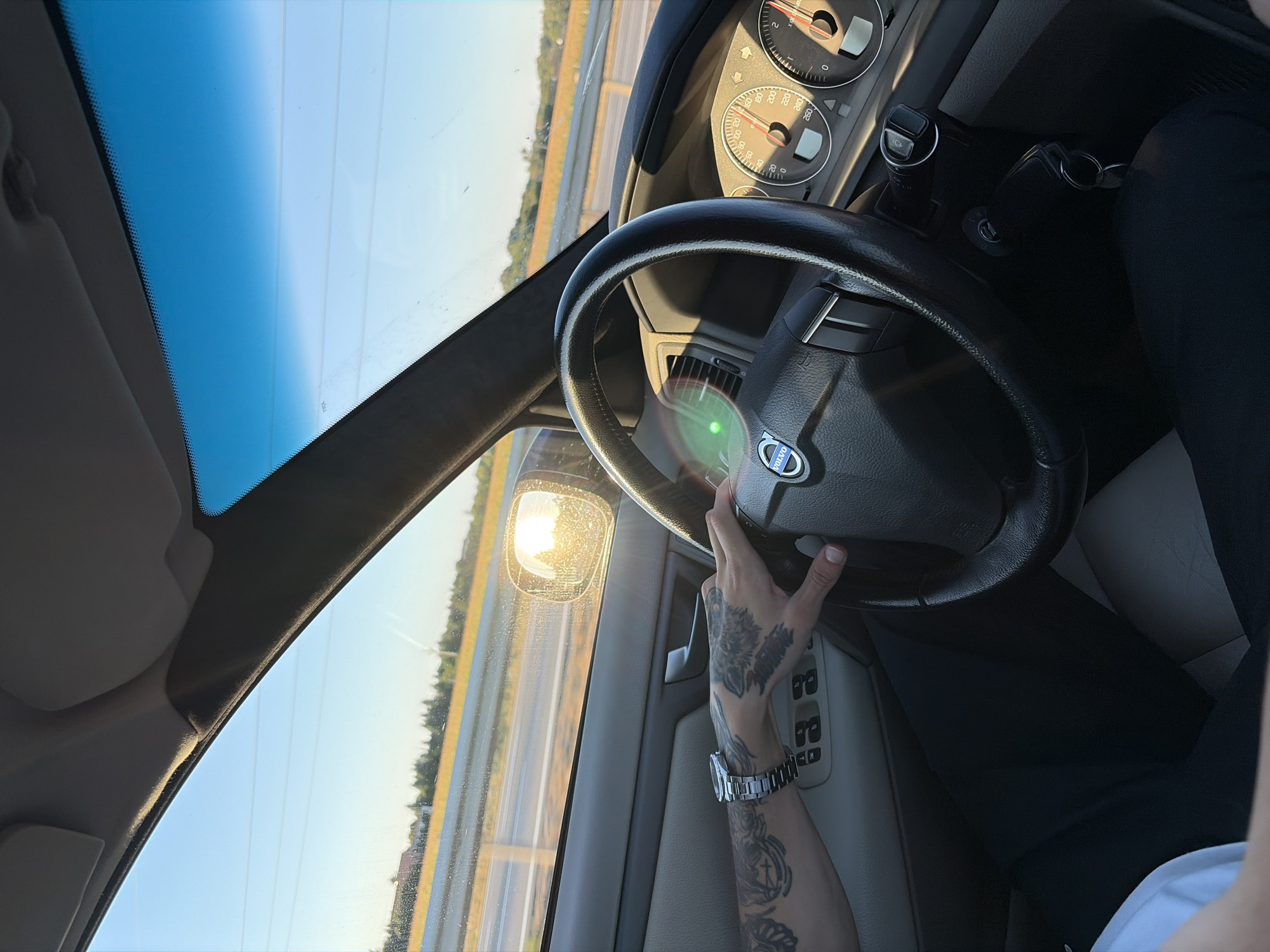 The image shows the interior of a car from the passenger's perspective, driving on a sunny day. A person's tattooed arm and hand are on the steering wheel, which has a Volvo logo in the center. The dashboard displays two gauges, one with a needle pointing to approximately 100 and the other to 0. The side mirror reflects the bright sun, casting a glare. Outside the car, a highway with yellow barriers and trees can be seen. The sky is clear blue with some distant power lines visible. The overall impression is of a scenic drive during daylight. The location is not specified as Pata, Slovakia.