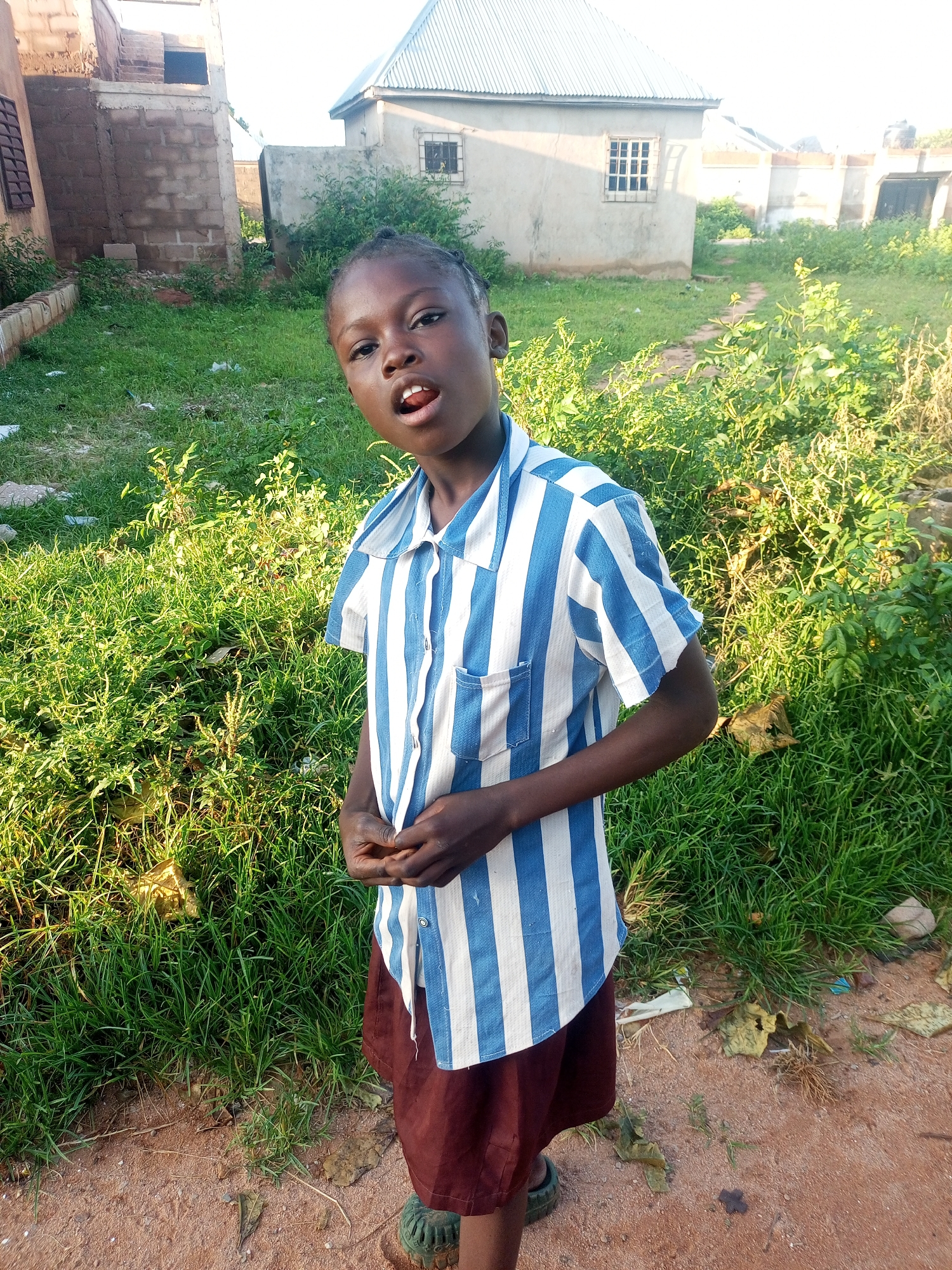 A young girl, appearing to be of African descent, stands in what seems to be an outdoor setting with some buildings and vegetation in the background. She is wearing a blue and white striped short-sleeved shirt and a dark red skirt. Her hands are clasped in front of her. The environment appears somewhat unkempt, with patches of grass and dirt. The lighting suggests it is daytime. The style of the buildings, particularly the one in the background with a corrugated metal roof, could be indicative of a rural or semi-urban area in Nigeria, possibly Tempure.