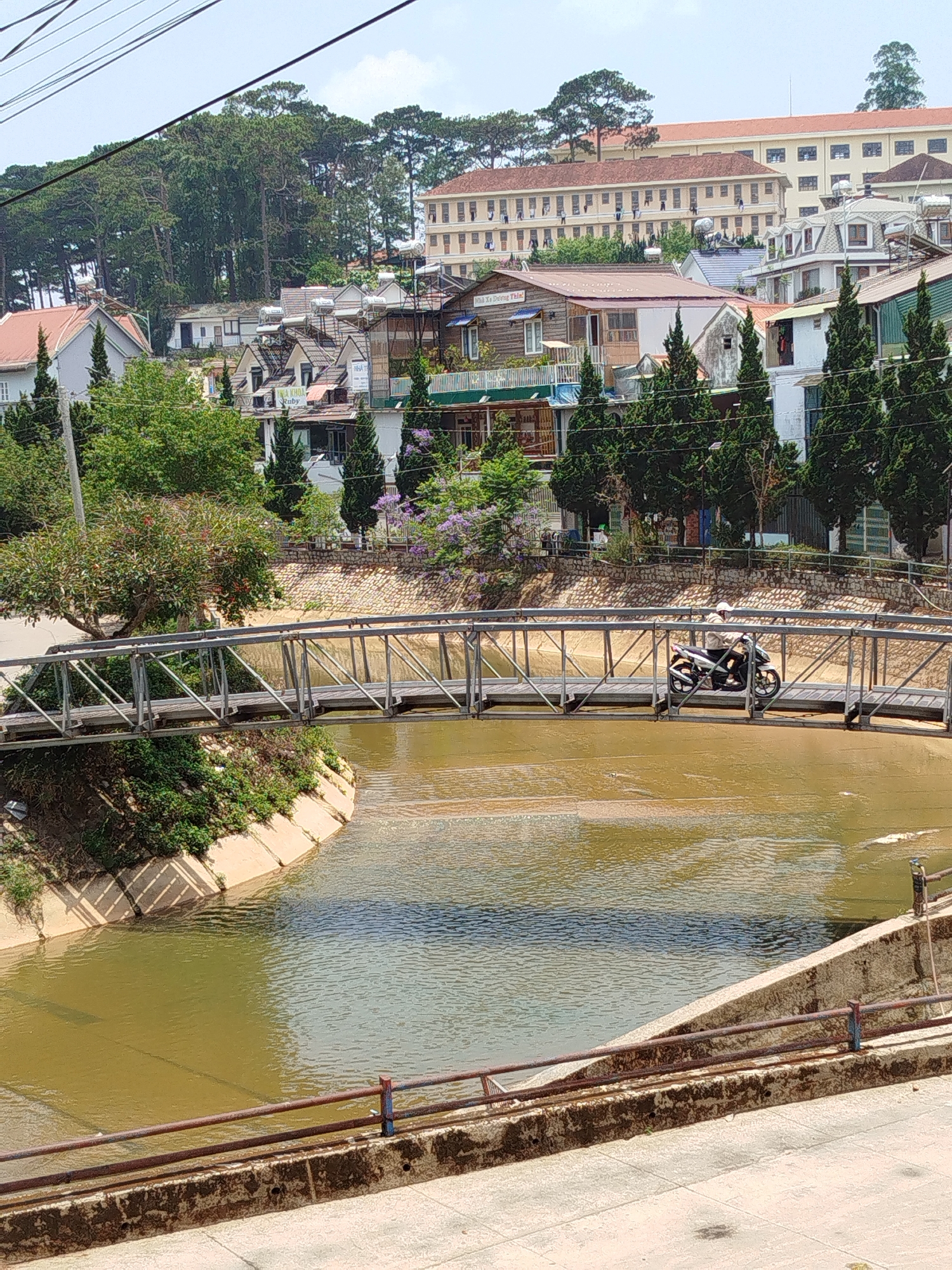A daytime outdoor shot of a steel bridge spanning a canal with murky, brown water, likely in the city of Cam Ly - Đà Lạt, Vietnam. A person on a moped crosses the bridge. The canal has concrete banks on either side. The bridge is supported by steel beams and has a wooden walkway. In the background, residential buildings are visible, with a large, pale building dominating the skyline. Trees surround the residential area. Overhead power lines are visible in the upper left corner. A rusted guardrail is in the foreground.