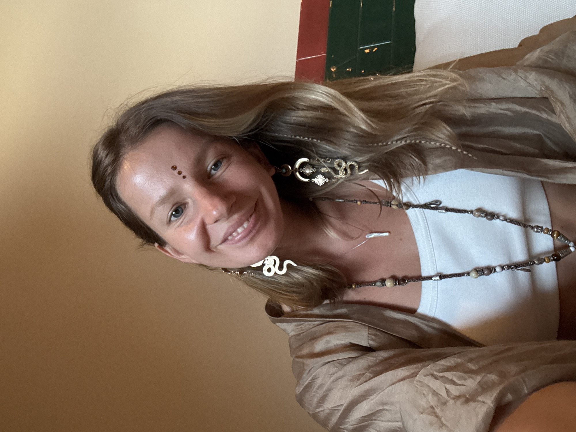 A young woman with blonde hair and blue eyes smiles into the camera. She has a henna design on her forehead, consisting of three small dots arranged in a triangle. She is wearing a white tank top and a silky, beige robe. Around her neck, she wears a long necklace made of various beads, including wood, stone, and metal. Her earrings are also notable: on her right ear, she has a dangling gold earring with a sun design, and on her left ear, a white snake-shaped earring. The background is a plain, warm-toned wall, with a glimpse of a green and red tiled structure in the upper right corner. The lighting suggests it might be daytime, possibly indoors, given the soft illumination. The overall mood is relaxed and friendly, with the woman appearing comfortable and at ease. There is no visible text in the image.