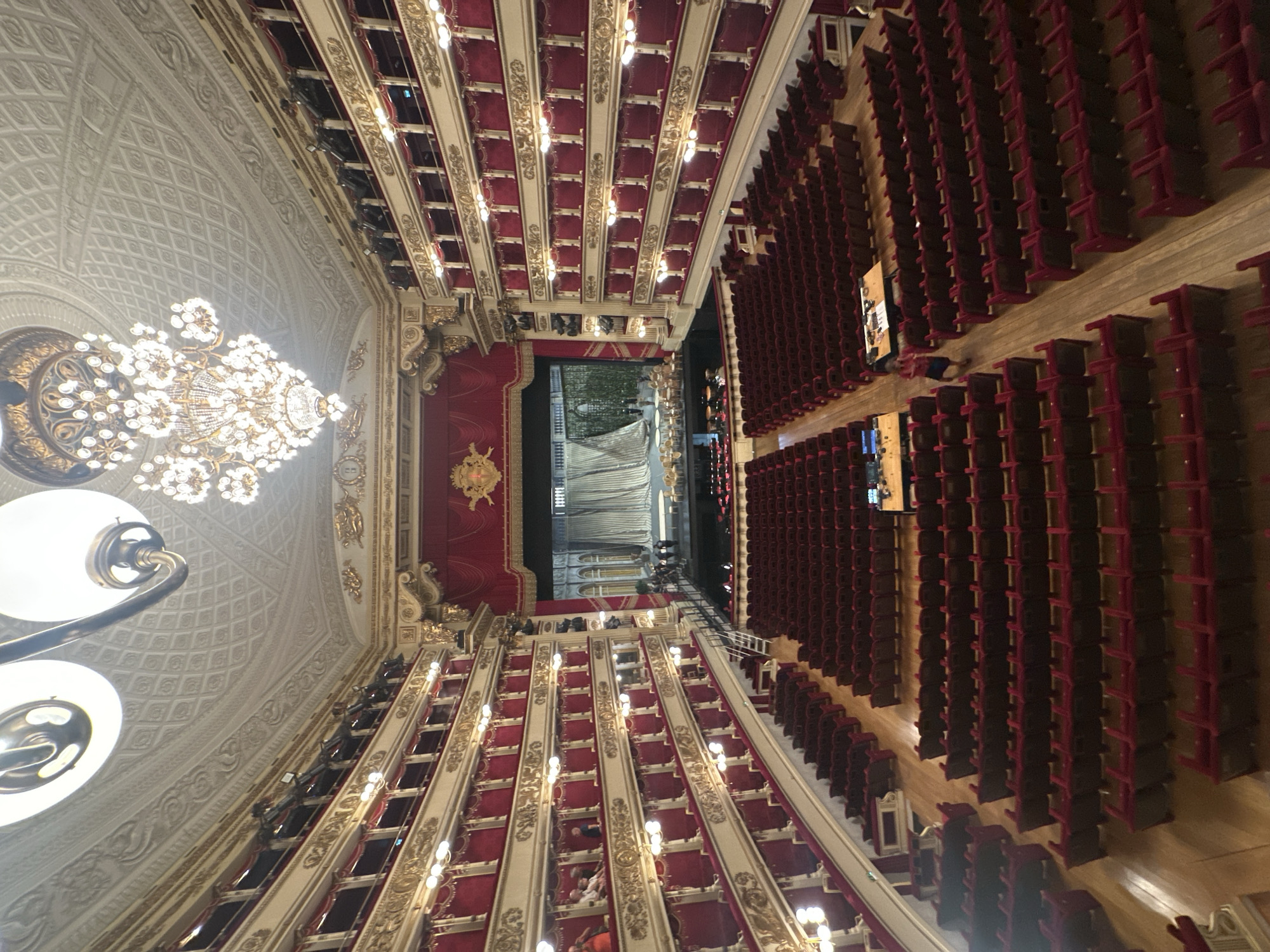 The image shows an overhead view of a grand theater or opera house. The scene is dominated by rows of plush, crimson theater seats, many of which are empty. The seats are arranged in tiers, with ornate gold detailing on the dividing structures between them. A large, sparkling chandelier hangs from the elaborately decorated ceiling on the left side of the frame. The ceiling itself features intricate white plasterwork with repeating geometric patterns and decorative molding.
On the right side of the image, more rows of red seats fill a large section of the theater. The floor appears to be made of polished wood. In the center of the image, the stage area is visible, with a red curtain and what looks like a grand staircase leading up to it. Some people are seated on the lower tiers of the theater, and a few individuals are visible on the stage area, suggesting the performance may not have started or has just ended. The lighting is a mix of bright overhead lights from the chandelier and stage area, and smaller, warm lights along the seating tiers. The overall impression is one of opulence and grandeur. There is no visible text in the image. The time of day and weather are not discernible.