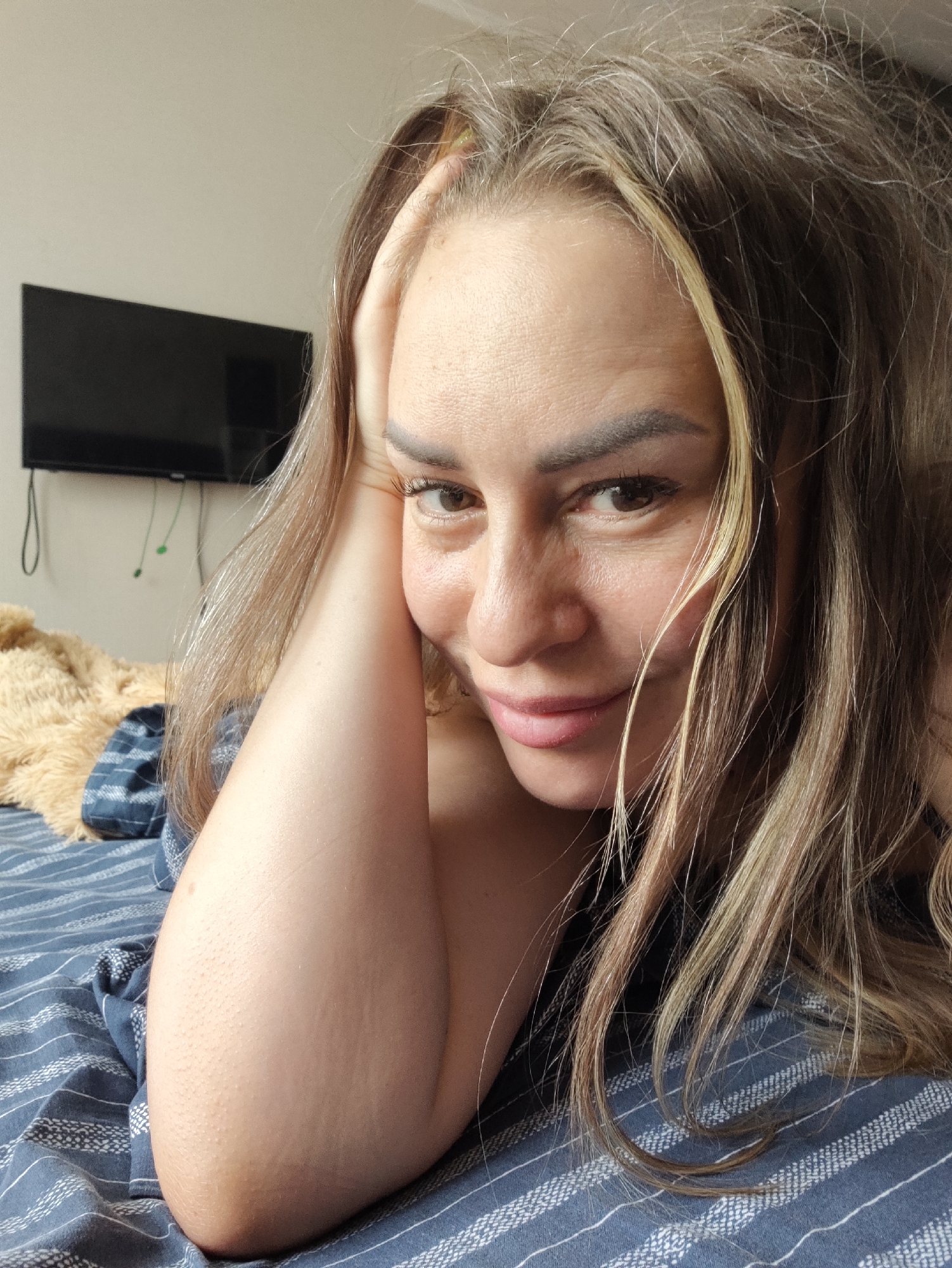The image shows a close-up of a woman with blonde and brown hair, lying on a bed with blue and white striped sheets. She is resting her head on her arm, and her left hand is touching her head. She is looking at the camera with a slight smile. In the background, there is a black television mounted on a light-colored wall and a tan-colored blanket. The scene appears to be indoors, in a bedroom. There is no information in the image to suggest it is located in Almaty, Kazakhstan.