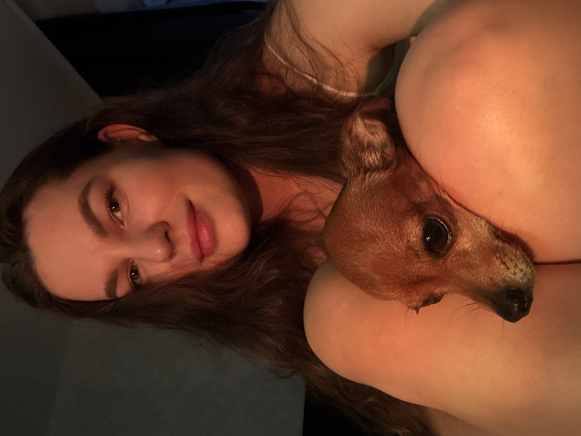 The image shows a young woman with long, dark, wavy hair, holding a small, tan dog in her lap. The woman is looking at the camera with a soft smile and her eyes are slightly closed. She is wearing a white tank top. The dog is resting its head on her thigh, with its eyes closed. The lighting is warm and soft, casting a golden glow on the scene. The background is dark and out of focus, with a hint of a dark blue wall on the right. The image appears to be taken indoors, in a casual setting. There is no visible indication of the location being in Chaikovsky, Russia.