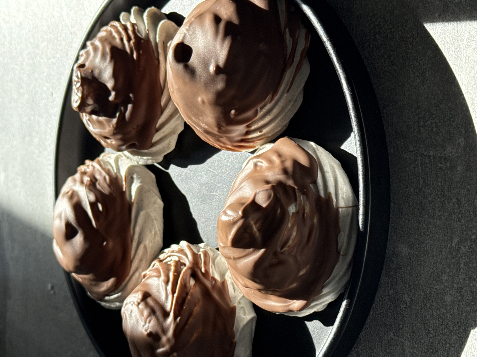 The image presents five individual confectionery items arranged on a dark, circular plate. Each item consists of a light-colored, possibly meringue or marshmallow-like, base with a textured, piped surface, almost entirely covered by a thick, smooth layer of milk chocolate. The chocolate coating shows slight irregularities and drips where it has solidified.
The plate is positioned on a gray, textured surface, appearing to be a countertop or table with a granular or speckled finish. Strong, directional lighting, likely natural, originates from the upper left of the frame, casting prominent, elongated shadows of the desserts and the plate onto the surface below. The background directly above the plate is a blurred, lighter gray tone. No people, actions, or interactions are depicted.
No specific geographical location, such as the city of Бордуки in Russia, can be determined from the visual information present in the image.