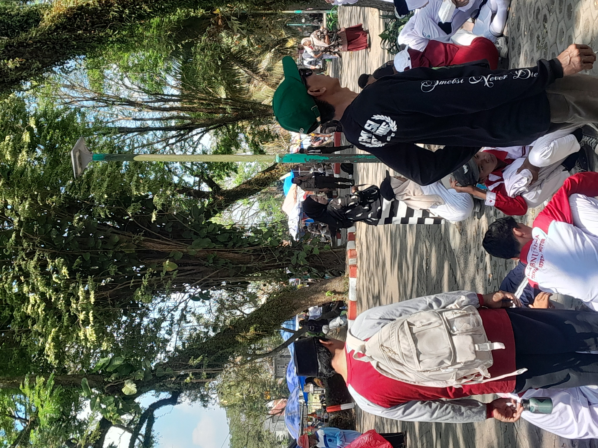 The image captures a daytime scene in what appears to be a public park or outdoor gathering space in Genteng, Indonesia. In the foreground, a man wearing a black long-sleeved shirt with white lettering and a green baseball cap is facing away from the camera, looking to the right. To his left, another man in a red and gray long-sleeved shirt and a black cap is also facing away, holding a small green cylindrical object in his left hand and wearing a light-colored backpack.
Several other individuals are visible throughout the scene. A woman in a black hijab and patterned dress is seated to the left of the man in the red and gray shirt. Further back, a uniformed individual, possibly a police officer, stands near some tents and stalls. Other people are seated or standing in the background, some in what appear to be school uniforms.
The environment is characterized by lush greenery with large trees providing shade, suggesting a tropical climate. The ground is paved with interlocking stones. In the background, colorful tents and umbrellas are set up, indicating a possible market or event. The overall atmosphere appears to be relaxed and communal.