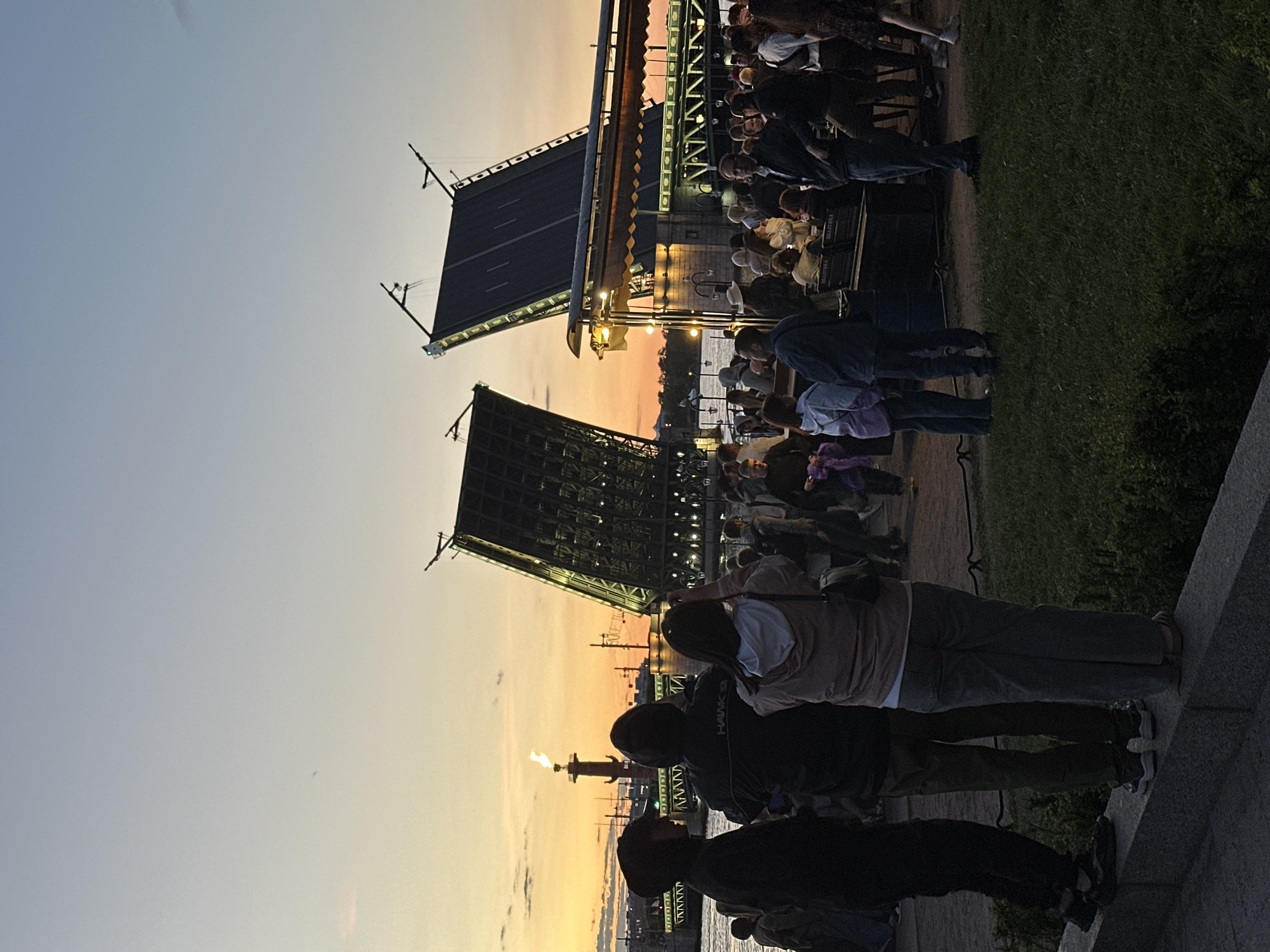 At dusk, a crowd of people stand on the embankment of the Neva River in Saint Petersburg, Russia, observing the Palace Bridge as it is drawn open for ships. The sky displays a gradient of colors, transitioning from a pale blue at the top to hues of orange and yellow near the horizon, indicating the setting sun. In the background, the Spit of Vasilyevsky Island is visible, marked by the Rostral Column with a lit torch. The Palace Bridge, a prominent architectural landmark, is elevated in two sections, revealing its intricate mechanical structure. Numerous individuals are captured in various poses, some facing the bridge, others looking at the sky or engaging in conversation. The foreground features a strip of grass and a stone ledge where some people are perched. The overall scene conveys a sense of communal enjoyment and admiration for a significant city event.