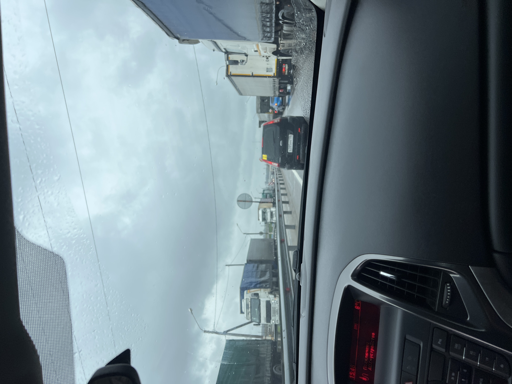 The image depicts a view from the interior of a car, looking through a rain-splattered windshield at a traffic jam in Rogachevka, Russia. The sky is predominantly grey and overcast, indicating a dull or rainy day. Raindrops are visible on the windshield, while part of the car's dashboard is seen on the right, featuring an air vent and an infotainment screen.
The screen displays the time "15:48" and an external temperature of "8°C". Cyrillic text on the screen reads "Подключено AUX-устройство" (Connected AUX device).
Outside the car, the road ahead is congested with heavy vehicles, mainly large trucks. Directly in front of the viewer's car is a black SUV or car with the Russian license plate "P280HH 196", which corresponds to the Sverdlovsk Oblast region. A yellow sticker is also visible on its rear window. Further ahead, numerous other trucks, including white ones and some with blue tarpaulins, are visible in the queue. Utility poles, overhead wires, and streetlights line the road, and a circular road sign is partially visible between two trucks. A road barrier or guardrail runs along the right side of the road. The scene portrays a slow-moving or stationary traffic condition, likely on a highway or major route, under cool and damp weather.