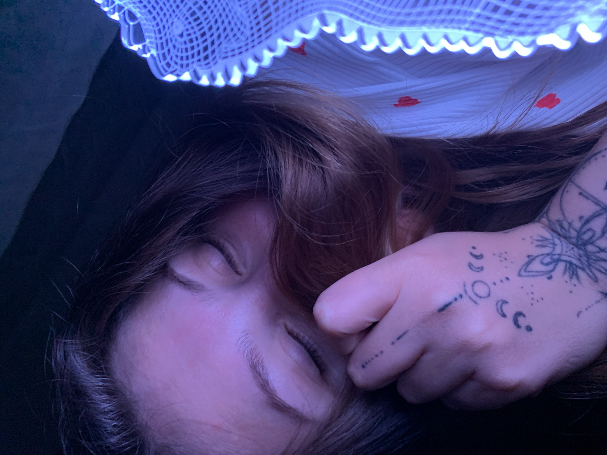 The image is a close-up, low-angle shot of a person's face and hand. The person is lying down with their eyes closed, their hair covering part of their face. They have tattoos on their hand and fingers, including celestial symbols and floral designs. The lighting is dim, with a purple hue cast over the left side of the image and a bright blue light illuminating the right side, possibly from a decorative light fixture or string lights. The person is wearing a white garment with small red heart patterns. The background is dark and indistinct. There is no discernible location in the city of Chaikovsky, Russia.