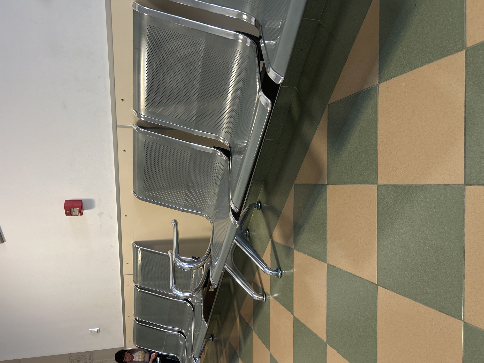 A low-angle, brightly lit interior shot presents a row of modern, metal public seating in a waiting area. The seating consists of multiple connected chairs, each with a perforated metal seat and backrest. Their silver frames and legs stand out against the tiled floor, which features a pattern of alternating light tan and green squares. The chairs align diagonally across the frame, casting shadows on the floor.
To the left of the seating is a blank, off-white wall with a red, rectangular fire alarm box mounted on it. Lower down on the wall, near the floor, are some small print signs or diagrams. Near the bottom of the frame, a person with short, dark hair can be partially seen, partially obscured by the last of the chairs. The location gives the impression of a clean, modern waiting area, possibly in a hospital or transportation hub.