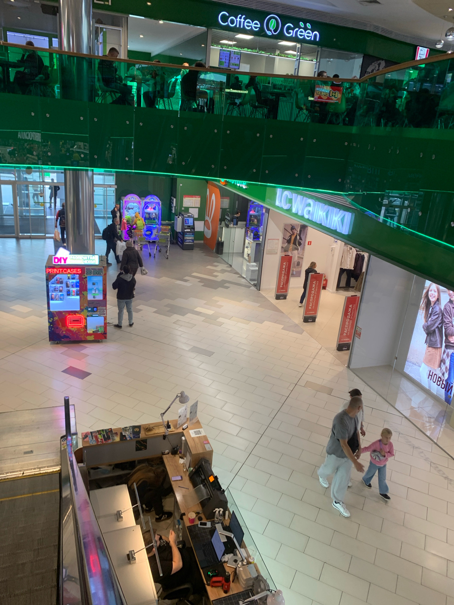 The image shows an interior view of a multi-level shopping mall, likely in Minsk, Belarus, given the presence of Cyrillic text on signage. The upper level features a "Coffee Green" establishment with patrons seated at tables. Below, a bright green railing delineates the levels. On the lower level, a "LC Waikiki" store is visible. In the foreground on the lower level, a man and a young girl are walking, with the man holding the girl's hand. To the left, a vending machine labeled "DIY PRINTCASES 3D BYN" is present, with a person standing near it. Further into the mall, there are other shoppers and store entrances. A kiosk with a computer setup and an escalator are also visible. The mall has a modern design with light-colored tiled flooring and glass railings.