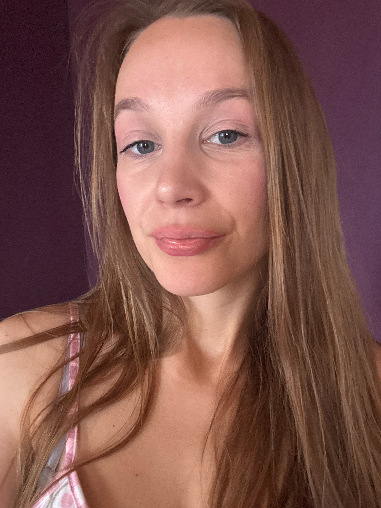 A close-up, head and shoulders selfie of a woman with long, light brown hair is shown against a purple background. She has blue eyes, fair skin with pink blush on her cheeks, and light pink lipstick. She is wearing a pink and white patterned top with thin straps. The background appears to be a wall. The location is not specified in the image or any accompanying data.