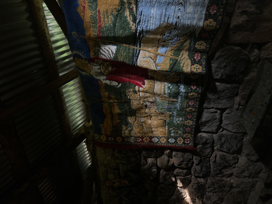 A tapestry hangs against a wall of rough, gray stones. The tapestry features a complex design with muted blues, greens, tans, and red. A border of repeating floral patterns runs along the edge. Light peeks through the stonework, illuminating parts of the wall. To the left, a corrugated metal wall is visible with sunlight filtering through. The overall scene is dimly lit.
