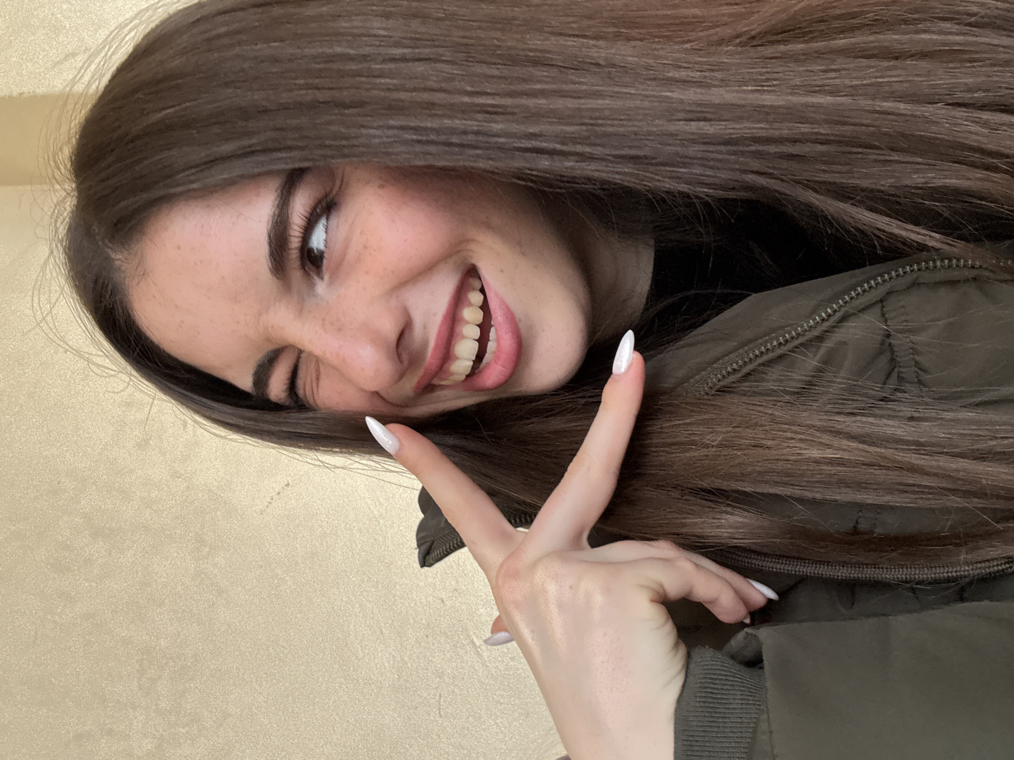 A young woman with long brown hair is smiling and winking at the camera while making a peace sign with her right hand. She is wearing a dark green jacket, and the background is a neutral color. The location is not identifiable as Plovdiv, Bulgaria, based on the image.