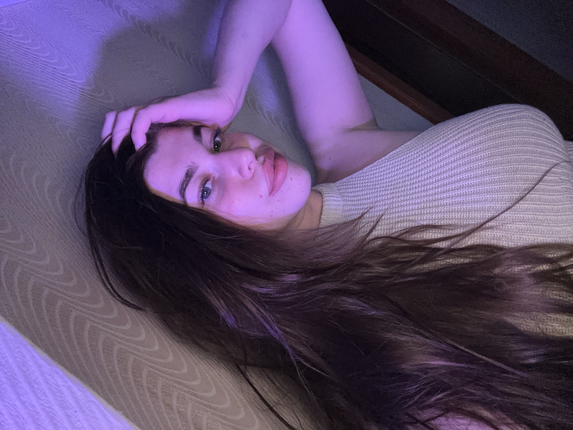 The media file is a close-up, high-angle shot of a young woman lying on her back. She has long, dark brown hair that cascades around her face and down her shoulder. Her eyes are open and looking towards the camera. She has light-colored skin with a few freckles visible on her cheeks and nose. Her lips are full and slightly parted, with a hint of pink lipstick.
The woman is wearing a light-colored, knitted sweater that appears to be off-the-shoulder. Her left arm is bent, and her hand is resting on the back of her head, near her hair. The lighting in the image is dim, with a prominent purple hue casting a soft glow on her skin and hair. This purple light appears to be coming from a light source to her left, illuminating a textured surface in the background. The background also includes a darker, wooden structure, possibly part of a roof or attic space, as well as a textured fabric, possibly a couch or bed.
The overall mood of the image is intimate and relaxed. The dim lighting and the woman's pose suggest a quiet, personal moment. The location context provided is Мещёры, Russia, though no specific landmarks are visible to confirm this. The time of day is likely evening or night due to the low lighting and artificial purple glow. There is no visible text in the image.