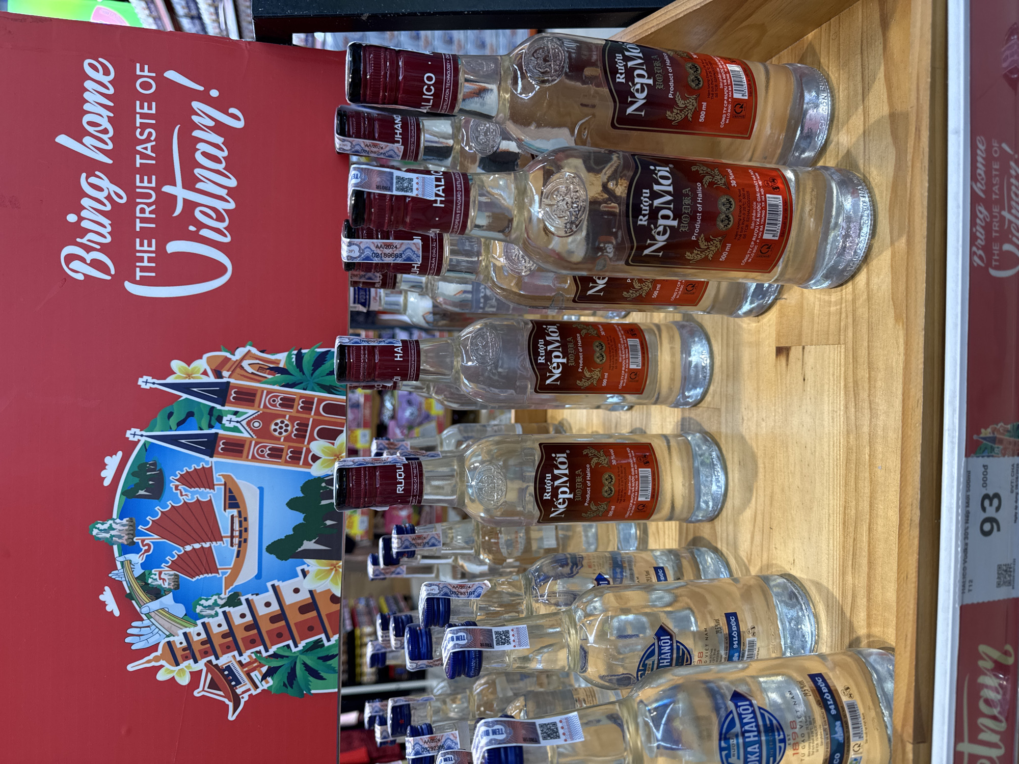 The image shows a display of Vietnamese vodka bottles on a wooden shelf. The bottles are clear glass with red and white labels. The labels indicate the brand is "Nếp Mới" and the product is vodka. Some bottles also say "Product of Halico". The bottles are arranged in multiple rows. Behind the bottles, a red banner is visible with text that reads "Bring home THE TRUE TASTE OF Vietnam!". The banner also features a colorful illustration of Vietnamese landmarks and scenes. The lighting is bright and appears to be from overhead store lights. The image is taken from a slightly elevated angle, looking down at the bottles. The background is blurred, showing shelves with more products and some reflections. There are no people visible in the image. The context suggests this is a retail environment, likely a supermarket or liquor store. The image does not provide specific information about the location being in the city of Đà Nẵng, Vietnam, as the text and imagery are general to Vietnam.