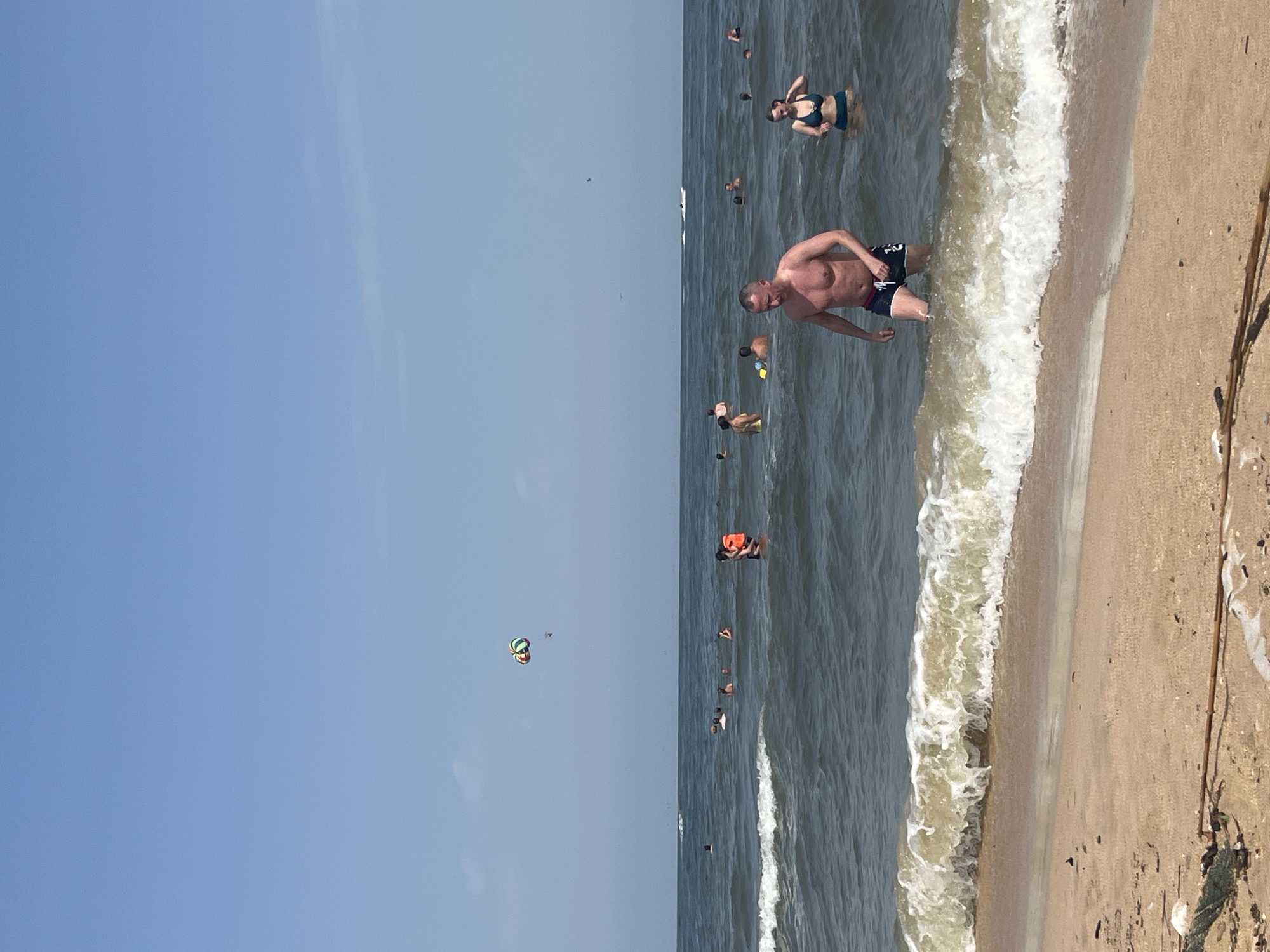 In Golubitskaya, Russia, a sunny day at the beach is depicted with a clear blue sky and calm ocean. The foreground shows a sandy beach with some debris. The water is filled with people swimming and enjoying themselves. In the middle ground, a man with a muscular physique is wading out of the water, wearing dark blue swim trunks. To his right, a woman in a blue bikini is also in the water. Further out, several other individuals are scattered across the sea. In the distance, a colorful parachute can be seen in the sky, suggesting a parasailing activity. A white boat is also visible on the horizon. The overall scene conveys a relaxed and recreational atmosphere typical of a summer beach outing.