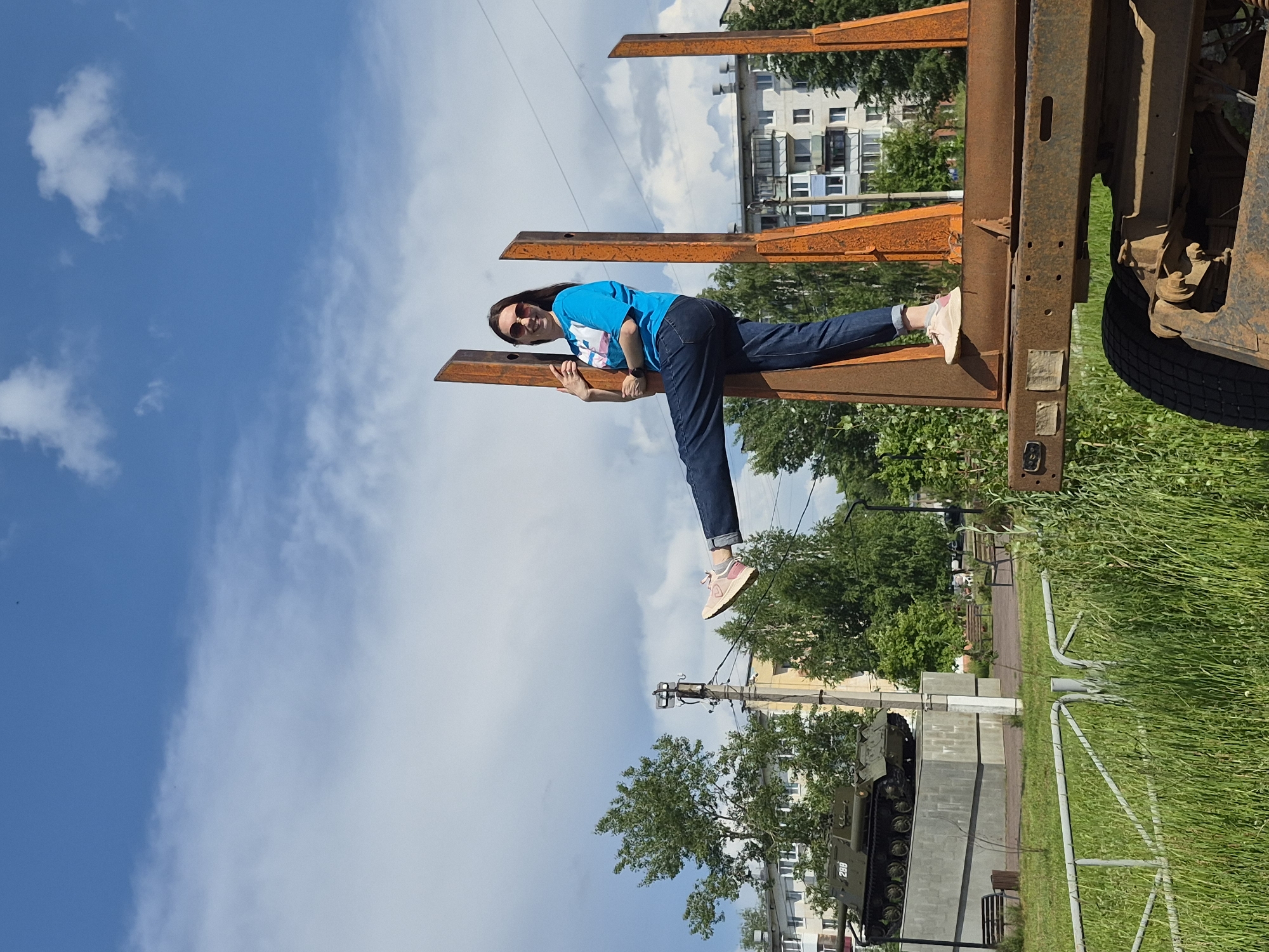 A woman in a blue t-shirt and jeans is posing on a rusty metal structure, possibly part of a trailer or platform, in Chaikovsky, Russia. Her right leg is extended forward, and she is holding onto a vertical metal beam with her left hand. She is wearing sunglasses and pink sneakers. In the background, there is a military tank on display on a concrete pedestal. Beyond the tank, there are trees, apartment buildings, and a blue sky with scattered white clouds. The foreground consists of tall grass and a metal fence.