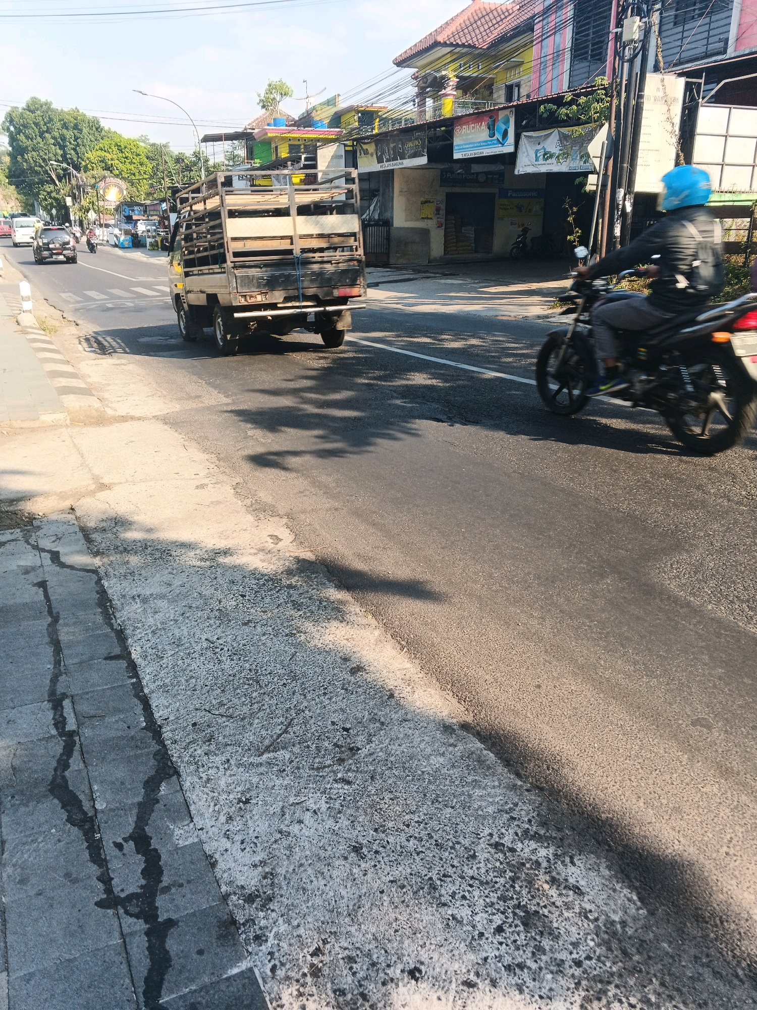 In Ungaran, Indonesia, a light beige truck with a metal cage on its bed drives down a road with a pedestrian walkway on the left. The truck is in motion, its rear visible to the camera. To the right of the truck, a person wearing a blue helmet and a dark jacket rides a black motorcycle, also moving forward. The road is asphalt and appears to be a multi-lane street with some traffic further down the road. Buildings line the right side of the street, some with colorful facades and signage in Indonesian. Trees are visible on the left side of the road in the distance. The sun casts long shadows across the road and walkway, indicating it is likely morning or late afternoon. The walkway is made of concrete tiles and has some water stains or dark marks.