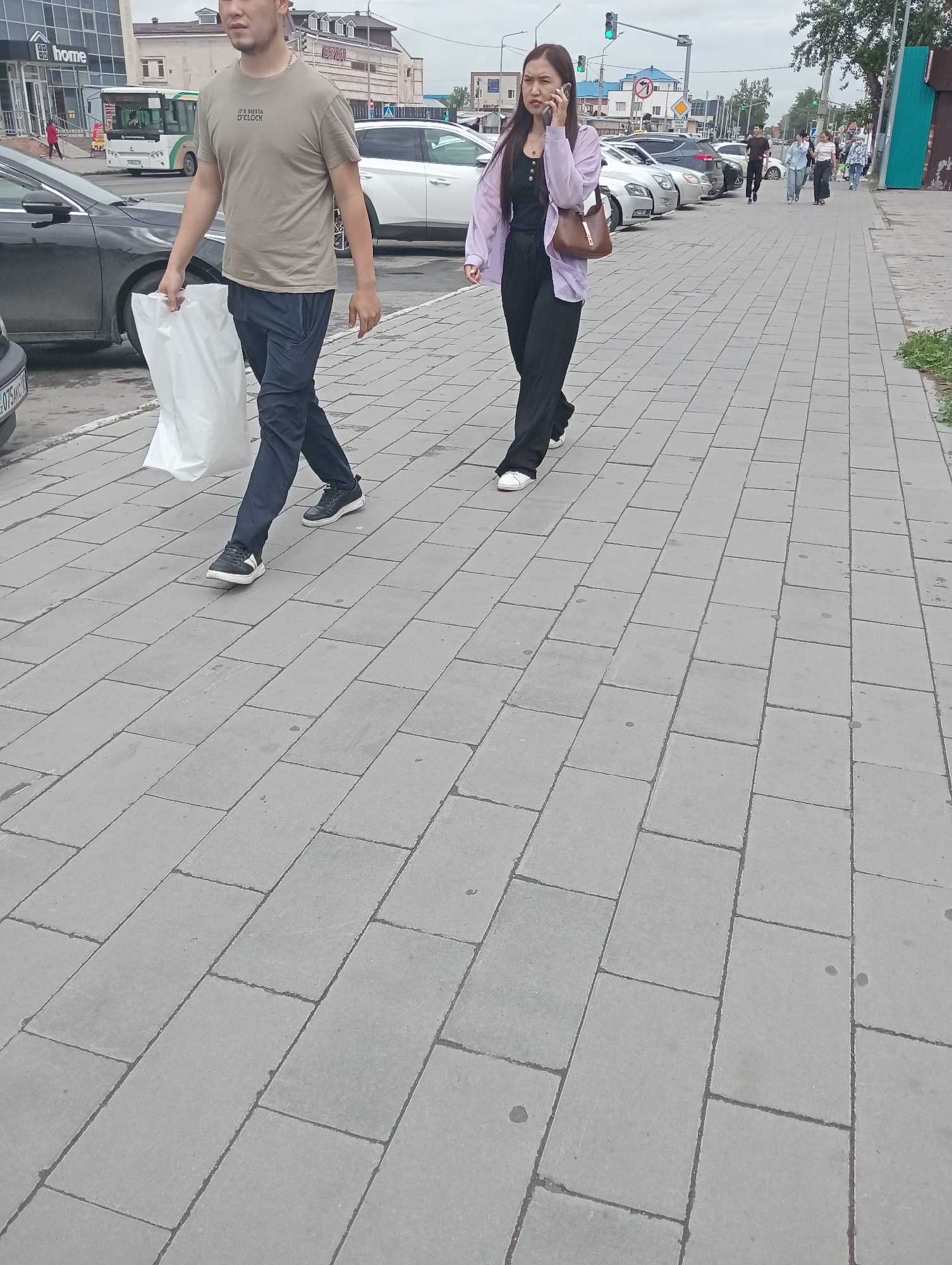 This image shows a street scene in Kostanay, Kazakhstan. Two people are walking on a paved sidewalk. In the foreground, a man is walking to the left, carrying a white shopping bag. He is wearing a light brown t-shirt and dark pants. Behind him, a woman is walking to the right, talking on her phone. She is wearing a black outfit with a light purple open cardigan and carrying a brown purse.
In the background, several cars are parked along the street. A white bus is visible on the left side of the image, and buildings line the opposite side of the street. Traffic lights are also visible, with a green light illuminated. The weather appears to be overcast, suggesting it could be daytime with cloudy skies. The overall scene depicts everyday urban life. Visible text includes "home" on a building in the background and text on the man's t-shirt which reads "IT'S SIX O'CLOCK".
