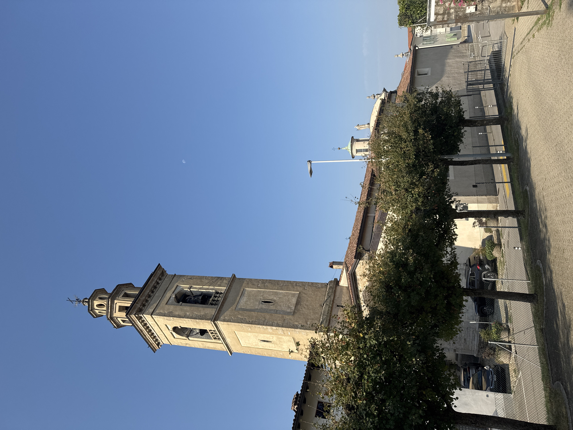 The image shows a view of the bell tower of the Church of San Giorgio in Balerna, Switzerland. The bell tower is made of stone and has a square base with arched openings for the bells. Above the bell chamber, there is an octagonal lantern with a cross on top. The church building is visible behind the bell tower, with a tiled roof and a small dome. In the foreground, there are several trees with green foliage, and a paved walkway. A metal fence separates the walkway from a parking area where several cars are parked. The sky is clear and blue, with a faint crescent moon visible. The overall scene is one of an outdoor urban environment with a historical building.