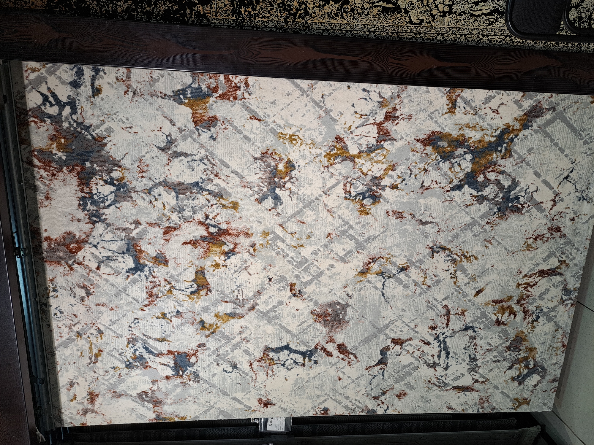 The image displays a large, abstract-patterned rug with a white, grey, blue, and rust color scheme, hanging vertically in a retail setting. The rug features a distressed, textured design with areas of what appear to be splotches and geometric lines. It is framed by dark wood trim on the right and a darker fabric on the left. In the background, to the right of the main rug, another black rug with a gold ornamental pattern is visible, partially obscured by the dark wood trim. The floor appears to be tiled. A small white tag with black text is attached to the upper left corner of the main rug. The scene is indoors, likely a rug store or showroom. The location is specified as Barnaul, Russia.