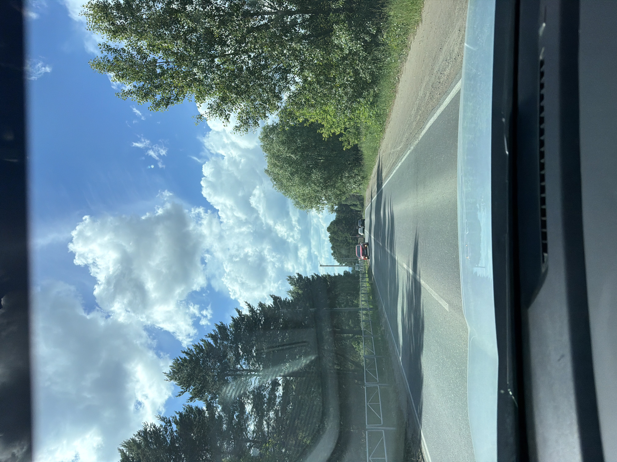 The image is a POV shot from a vehicle traveling on a paved road. The road is flanked by trees and grass, with a white metal fence on the left side. In the distance, several cars are visible on the road, including a red car. The sky is blue with scattered white clouds. The scene appears to be in a rural or suburban area. The image does not provide enough information to definitively pinpoint the location within the city of Chaikovsky, Russia.