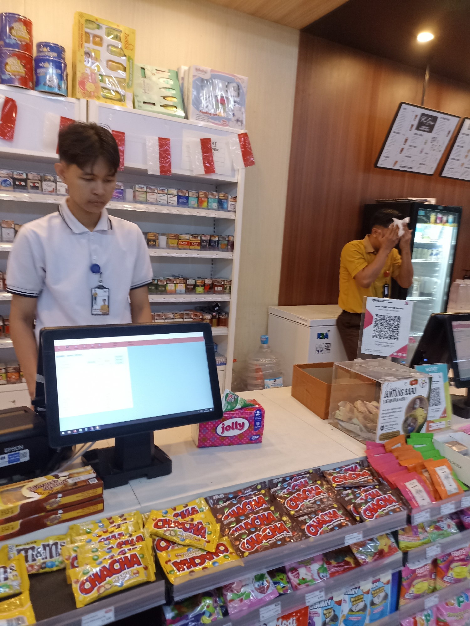 The image depicts the interior of a convenience store, likely located in Tompokersari, Indonesia, given the context. In the foreground, a young male cashier stands behind a point-of-sale system, which includes a monitor displaying a retail interface. To the right of the cashier, a second male employee, wearing a yellow shirt, is seen wiping his face with a white tissue. He appears to be sweating, possibly due to the ambient temperature or the operation of nearby refrigerators.
The store shelves are stocked with a variety of retail products, including snacks, candy, and possibly beverages. Packaged candies, such as "Chacha" and "M&M's," are prominently displayed at the checkout counter. A large water cooler is visible in the background, along with a freezer unit and a refrigerator stocked with bottled drinks. The overall environment suggests a typical retail setting, with goods arranged for customer purchase. The lighting appears to be a mix of overhead fixtures and ambient light. The arrangement of products and the presence of a point-of-sale system indicate a functional retail operation.