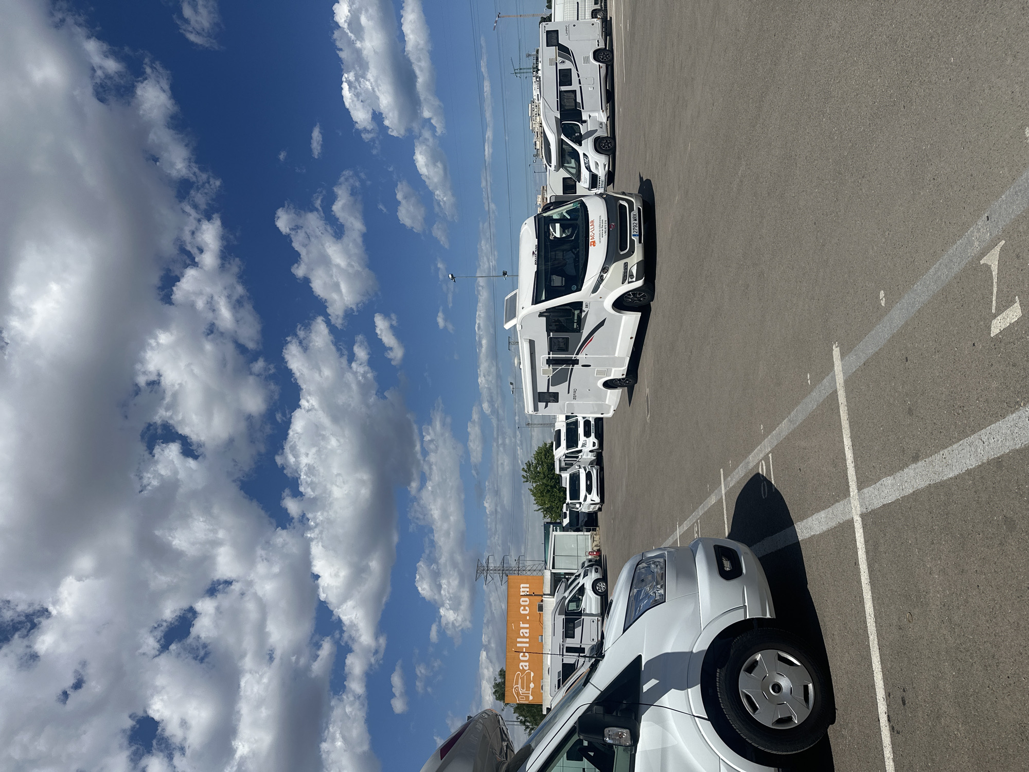 The image depicts a parking lot filled with recreational vehicles (RVs) in Quart de Poblet, Spain. The RVs are predominantly white. An orange billboard with "ac-llar.com" is visible in the background. The sky is blue with puffy white clouds. Power lines stretch across the horizon. The parking lot is marked with white lines, and the parking space numbers are visible. The image appears to have been taken on a sunny day.