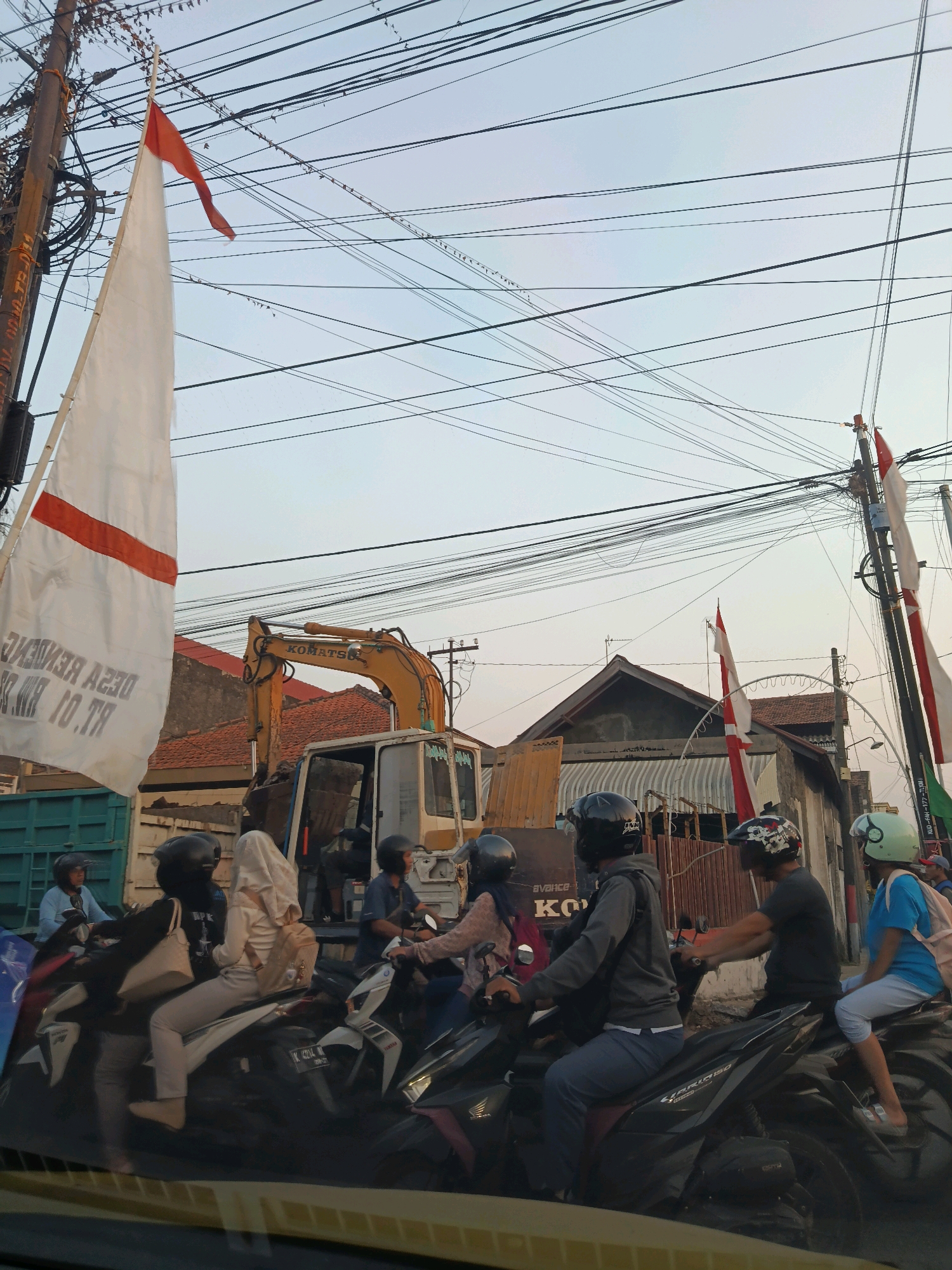 The image depicts a street scene in Burikan, Indonesia, characterized by a dense network of overhead wires. In the foreground, several people on motorcycles are stopped, possibly in traffic. The individuals are wearing helmets and casual clothing. Some are looking forward, while others appear to be interacting with each other. A large white flag with a red stripe and text is prominently displayed on the left, attached to a pole and a truck. In the background, an excavator is visible next to a building with a red-tiled roof. Other flags, also red and white, are seen on poles to the right. The overall environment suggests a busy urban or semi-urban setting.