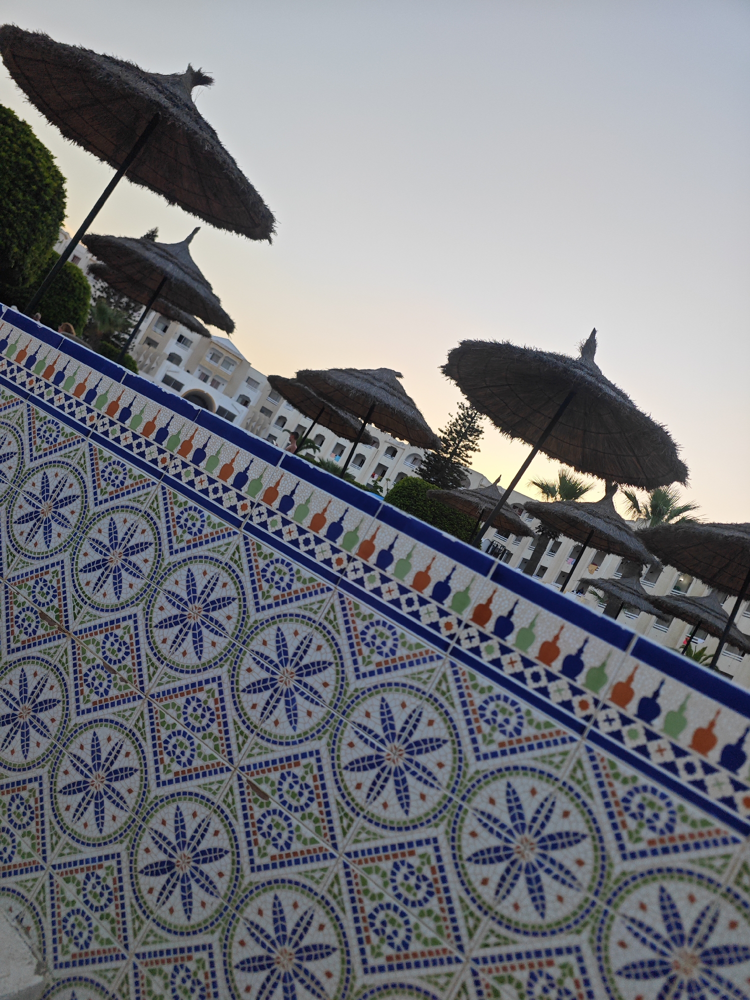 This is an angled shot of a tiled surface with a patterned design. Behind the tiled surface, there are several straw umbrellas standing in front of a white building. The sky is a pale, hazy color, suggesting either dawn or dusk. The building appears to be a hotel or resort in Mahdia, Tunisia, as indicated by the presence of multiple umbrellas and a building structure commonly found in tourist areas.