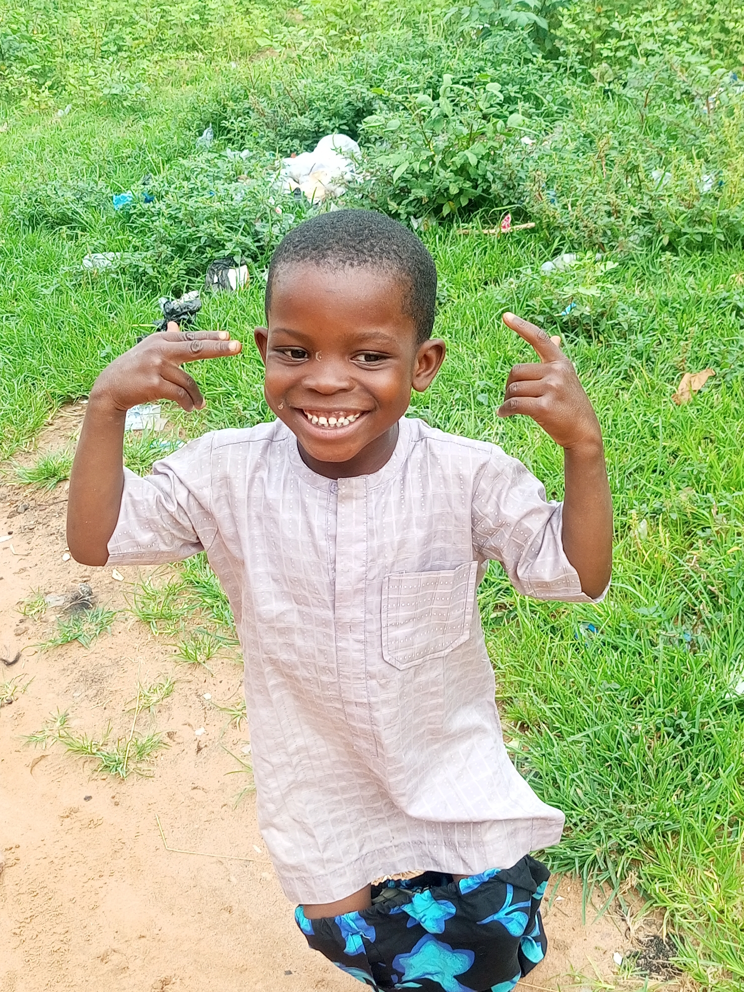 A young boy of West African descent, estimated to be between 5 and 8 years old, stands in an outdoor environment characterized by sparse sandy ground and overgrown green vegetation. The boy is wearing a light-colored, long-sleeved tunic with a subtle checkered pattern and a banded collar. Below the tunic, he is wearing black shorts adorned with a blue floral print. His skin tone is dark brown, and his hair is short and dark.
The boy is positioned centrally in the frame, facing slightly towards the viewer. He has a broad, toothy smile, revealing white teeth. His facial expression suggests happiness or playfulness. His arms are raised, with his index fingers pointing towards his temples, a gesture that could be interpreted as playful or mimicking.
The background consists of dense green grass and foliage, with some scattered debris, including white plastic bags, visible amongst the plants. The ground in front of the boy is sandy and dry, with a few sparse blades of grass. The lighting suggests it is daytime, possibly overcast, as there are no strong shadows. The scene appears to be in a rural or semi-rural setting within Tempure, Nigeria, likely an area with undeveloped land.