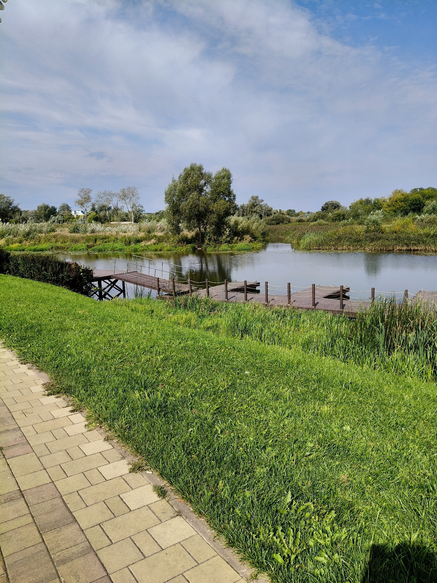 The image depicts a serene waterside scene with a paved walkway bordering a lush green lawn. Beyond the lawn, a wooden dock extends into a calm river, surrounded by dense vegetation and trees under a partly cloudy sky. The scene is tranquil and showcases a well-maintained outdoor area, likely a park or recreational spot. There are no people or animals visible in the image. The location in Naro-Fominsk, Russia, is implied to be a natural or landscaped area within the city.