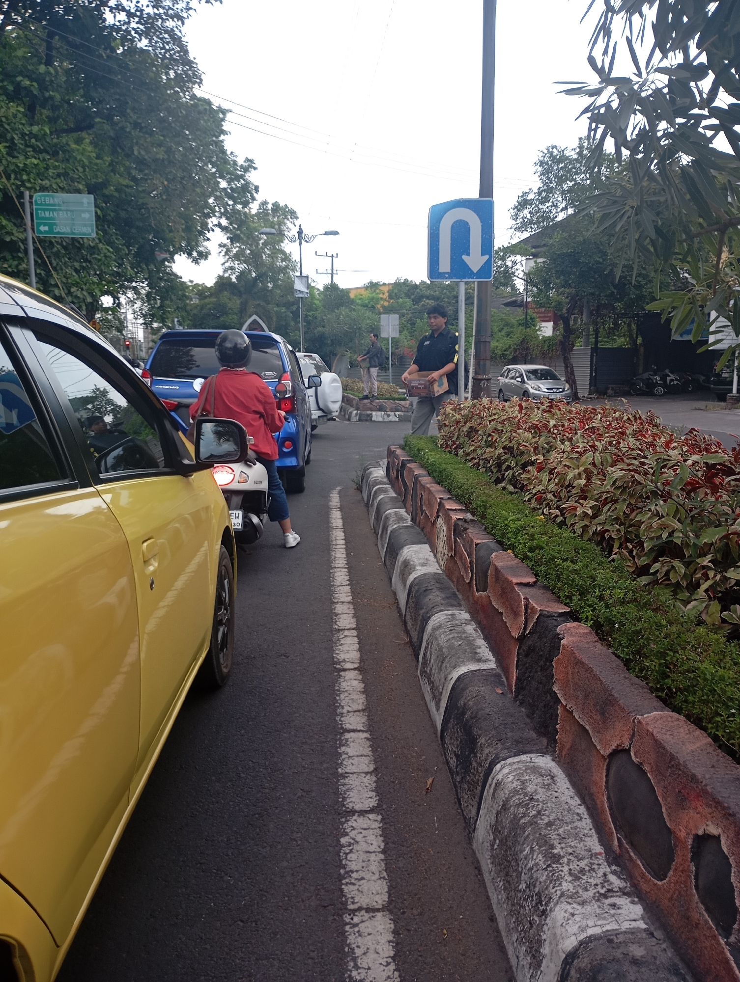 The image depicts a traffic scene on a road in Mataram, Indonesia, likely during the daytime due to the overcast sky. In the foreground, a yellow car is partially visible on the left side of the frame. Ahead of the yellow car, traffic is backed up, with a blue SUV and a white scooter in the middle of the lane. A person wearing a red jacket and blue jeans is riding the scooter, facing away from the camera and wearing a black helmet. To the right of the scooter, there is a median strip with decorative plants and a concrete barrier painted with black and white stripes. A man in a dark uniform, possibly a security guard, is standing on the sidewalk next to a U-turn traffic sign. He is holding a cardboard box. Further down the road, other vehicles and pedestrians are visible. The environment appears to be urban, with trees lining the street and buildings in the background.