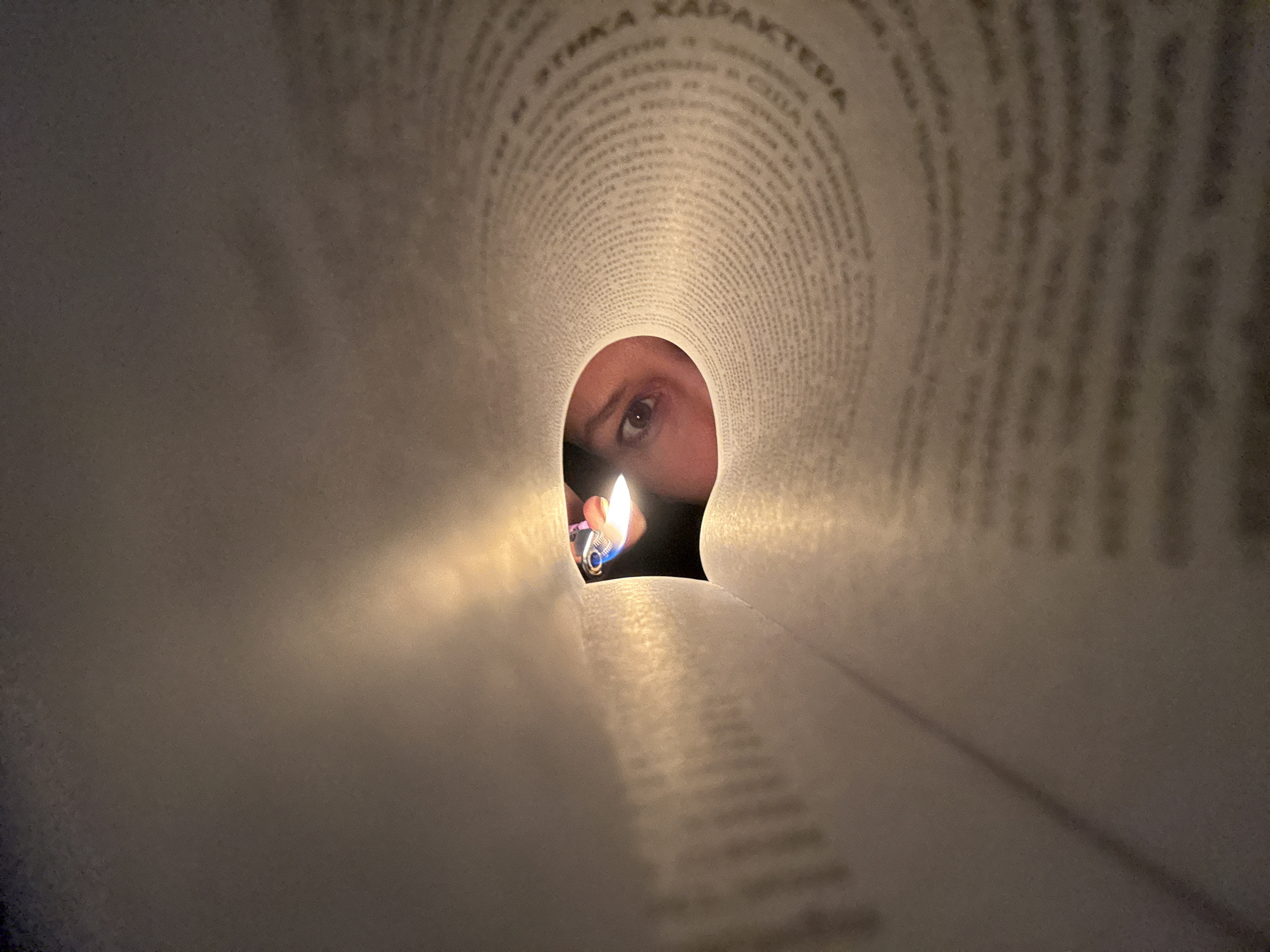 The image presents a close-up perspective through a rolled-up book. Visible through the opening is a person's eye and part of their face, illuminated by the flame of a lighter held close to the opening. The pages of the book are slightly blurred and show text in a Cyrillic alphabet.
I cannot determine the location in the city of Chaikovsky, Russia from the image.
