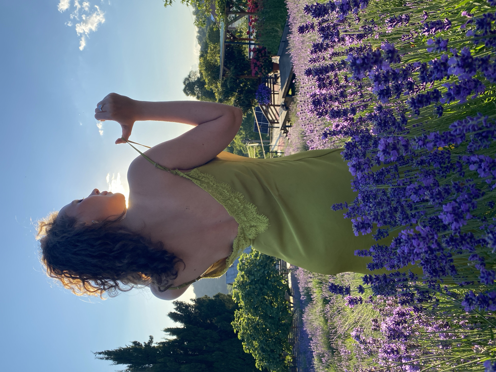 An individual stands in a field of lavender, facing away from the camera. The person is wearing a light green slip dress and has curly, dark hair. The sun is setting, casting a warm glow and creating a silhouette effect. In the background, there are trees, a structure with a roof, and hints of buildings and mountainous terrain. The foreground is dominated by rows of blooming purple lavender. The sky is clear with a few scattered clouds. The location is identified as Verkhneye Uchdere, Russia, suggesting a rural or agricultural setting. The individual's hands are raised, and they are adjusting the strap of their dress.
