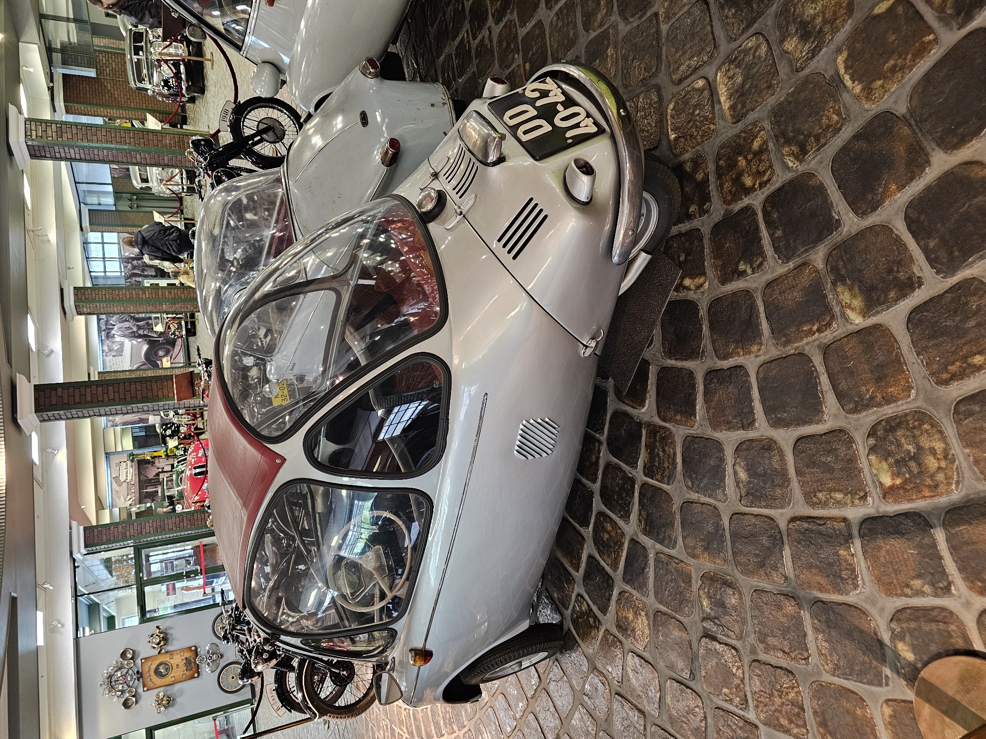 The image shows a light-colored vintage car, likely an Isetta or a similar microcar, parked on a cobblestone floor in what appears to be a museum or exhibition space. The license plate on the car reads "DD 40-42". Adjacent to it are other vintage vehicles and motorcycles. In the background, architectural details such as pillars, windows, and possibly a hallway can be seen. The city of Архангельское, Russia is not explicitly visible in the image, but it can be inferred based on metadata, if any. The overall lighting suggests an indoor setting with controlled lighting, possibly a museum display.