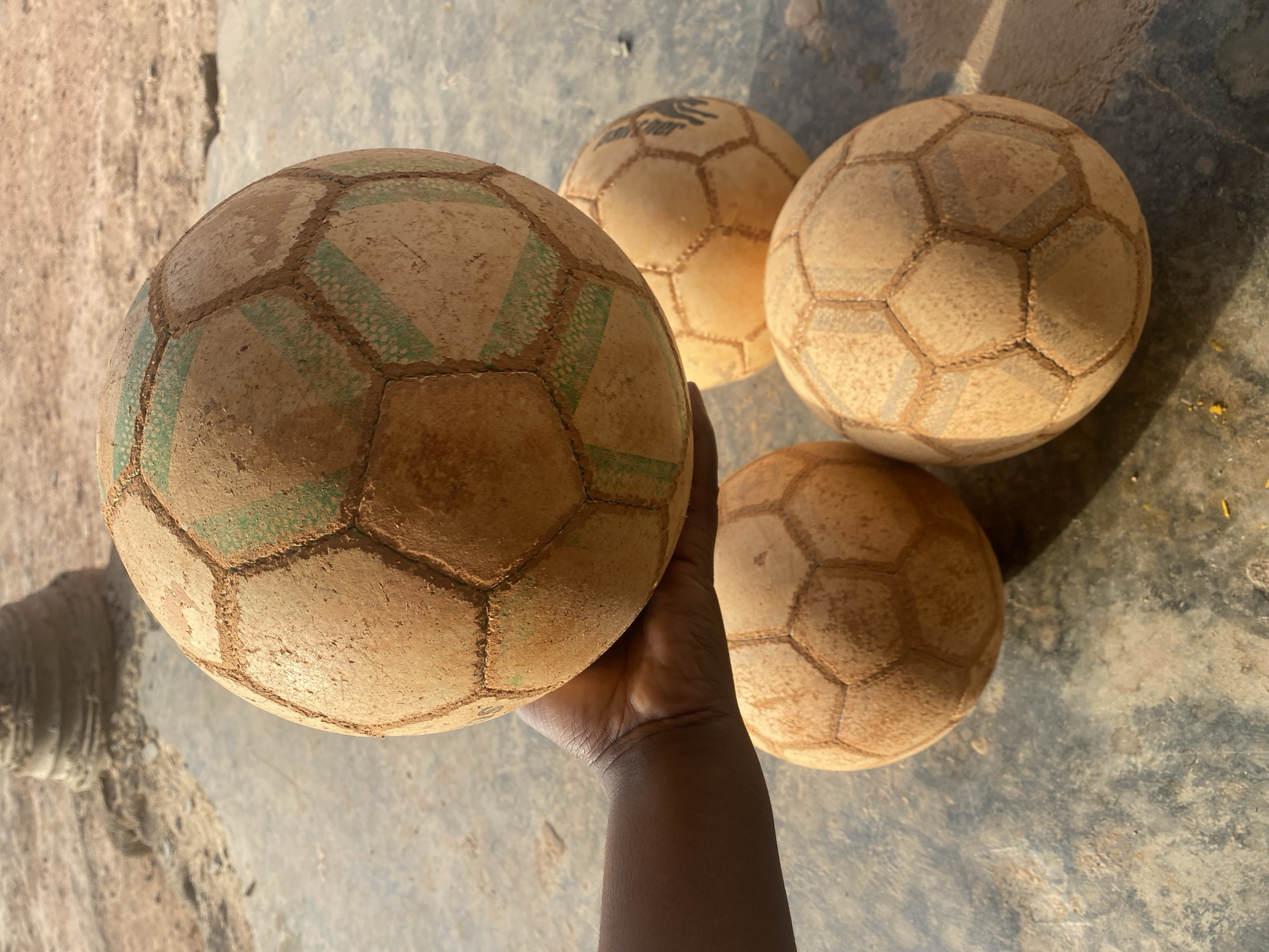 The image presents an overhead shot of four heavily used footballs (soccer balls) on a rough, earthy ground, with one ball being held by a dark-skinned hand. The scene is illuminated by bright daylight, casting distinct shadows.
In the immediate foreground, a large, dirt-covered football is prominently featured, held from below by a dark-skinned hand. The ball is a muted light brown or tan color, caked with dirt, which highlights the stitching lines that form its classic hexagonal and pentagonal panels. Some panels on this ball show remnants of faded green striped patterns, barely visible beneath the layer of grime. The hand itself is dark-skinned, gripping the ball firmly with fingers and palm visible, extending slightly into a lower forearm.
Behind the held ball, three other footballs are scattered on the rough ground surface. These balls also exhibit significant wear and tear, appearing equally dirty and scuffed, predominantly in various shades of light brown and tan. One of these background balls, positioned centrally behind the held ball, clearly displays a faded black logo on one of its panels, which reads "Derbystar" above a stylized hand or star emblem. The other two balls are less distinct but show similar signs of heavy use.
The ground beneath the balls is uneven and appears to be a mix of dirt, concrete, or a similar worn outdoor surface. It features varying textures and patches of grey, brown, and yellowish hues, consistent with a dry, dusty environment. Long shadows cast by the hand and balls indicate that the photograph was taken under direct, bright sunlight, suggesting a midday or early afternoon setting. The overall impression conveyed is one of well-loved sporting equipment, perhaps from a game played in an unpaved or informal outdoor area.