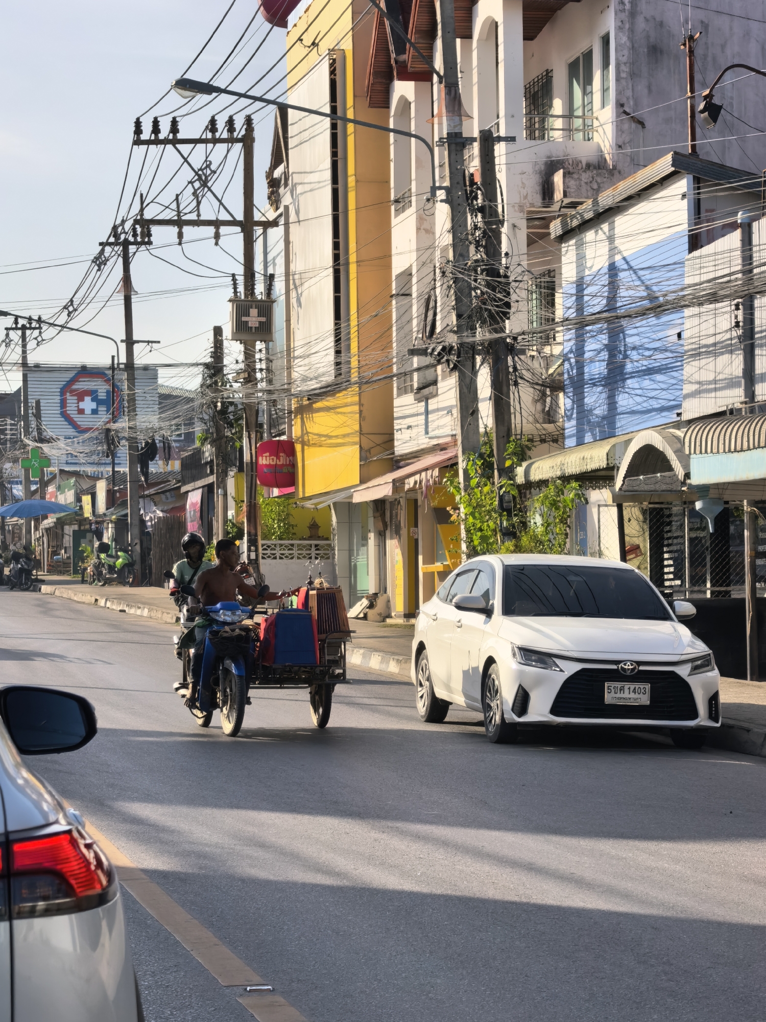 In the city of Ko Samui District, Thailand, on a street with utility poles and overhead wires, a white sedan is parked on the right side of the road, facing left. In the center of the image, two individuals are riding a blue motorcycle with a sidecar. The motorcycle is pulling a cart filled with blue and red containers, as well as a woven basket. The motorcycle and cart are moving away from the viewer. Buildings line the street on both sides, featuring a variety of colors and architectural styles. The road surface is asphalt, with a yellow line marking the lane. The sky is clear and blue.
