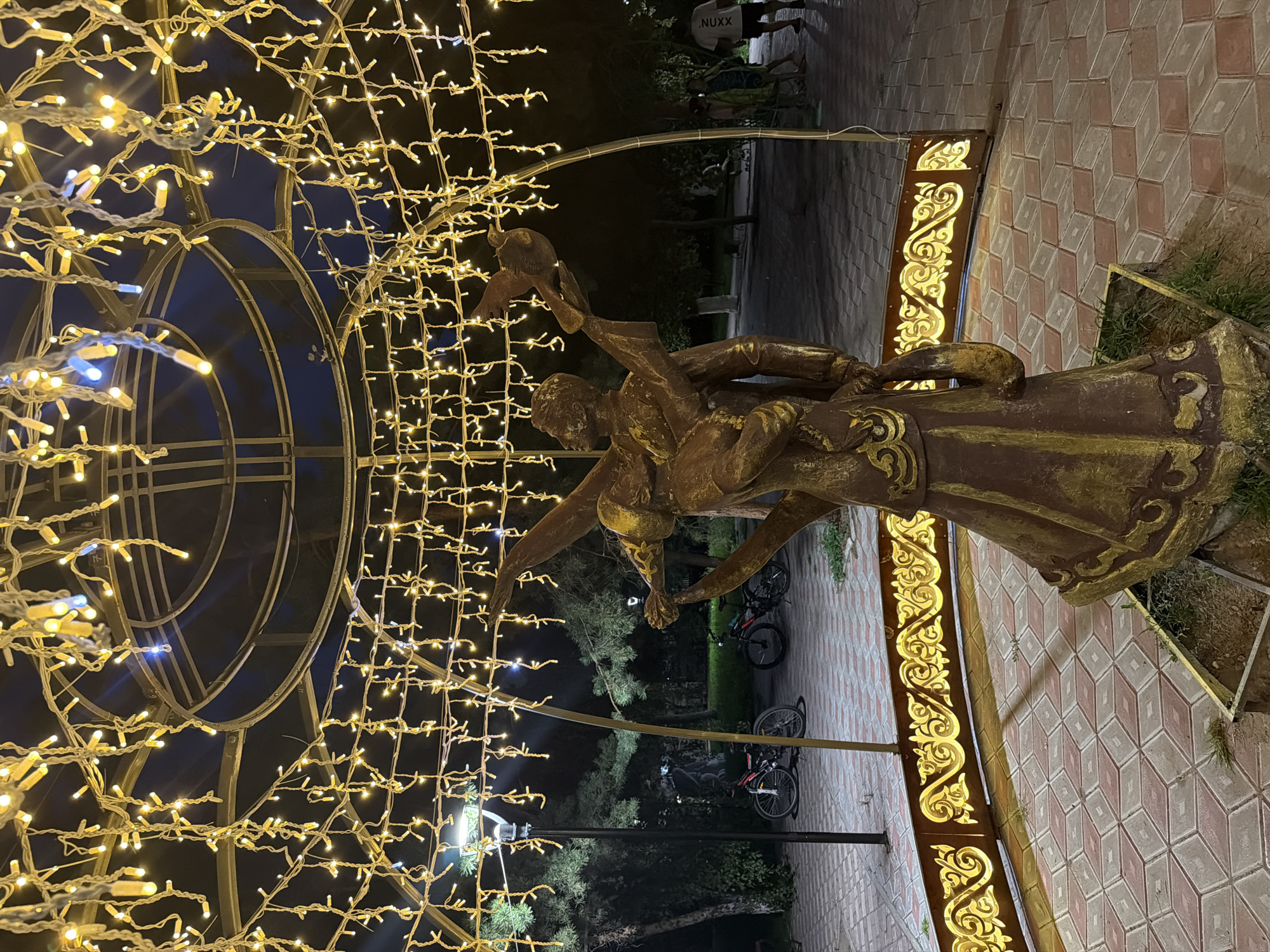 The image shows a statue of a couple in a park at night. The statue is illuminated by strings of yellow lights that hang from a circular structure above. The couple is depicted in a dance-like pose, with the man holding a bird in his outstretched hand. The park has paved ground with a red and grey diamond pattern. In the background, there are trees, a lamppost, and two bicycles. A few people are visible in the distance. The overall scene suggests a festive or romantic atmosphere. This is likely a public park in the city of Pavlodar, Kazakhstan, decorated for an evening event or celebration.