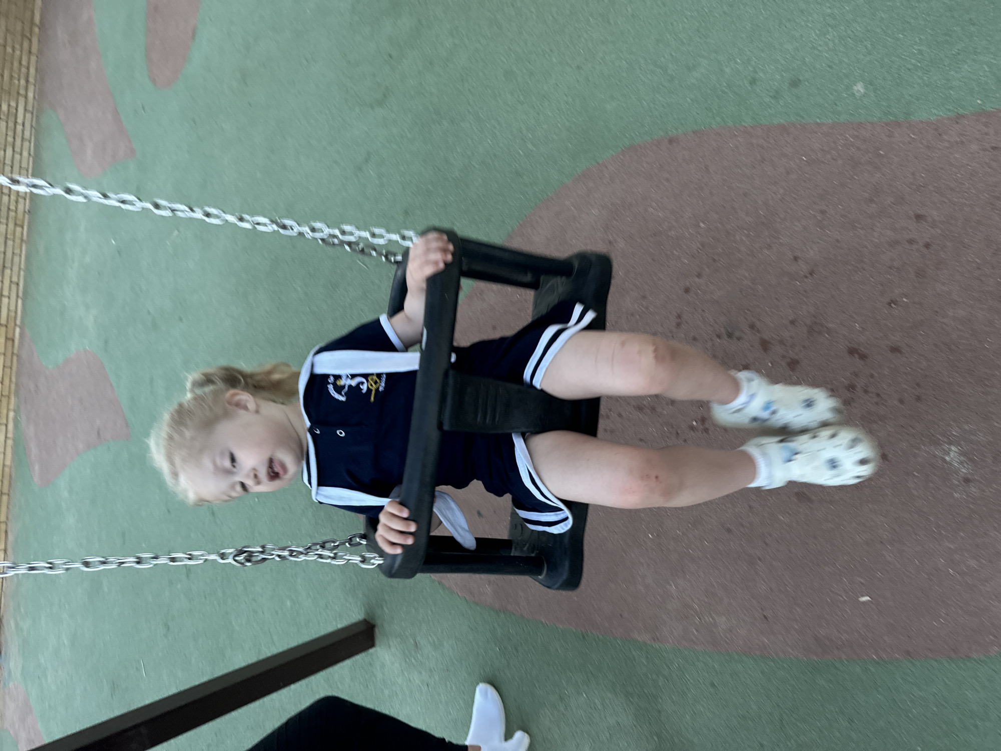 A young child with blonde hair pulled into a ponytail is swinging on a dark-colored swing. The child is wearing a navy blue sailor-style dress with white trim and white socks with white clogs. The child's knees are visible and appear to have minor abrasions. The swing is suspended by metal chains from a dark brown swing set frame. The ground beneath the swing is a rubberized surface with patches of green and brown. In the background, a brick wall is visible. To the left of the swing set, a person's legs and feet are visible, wearing black pants and white sneakers. The scene appears to be a playground.
This image was taken in the city of Sant Feliu De Guíxols, Spain.