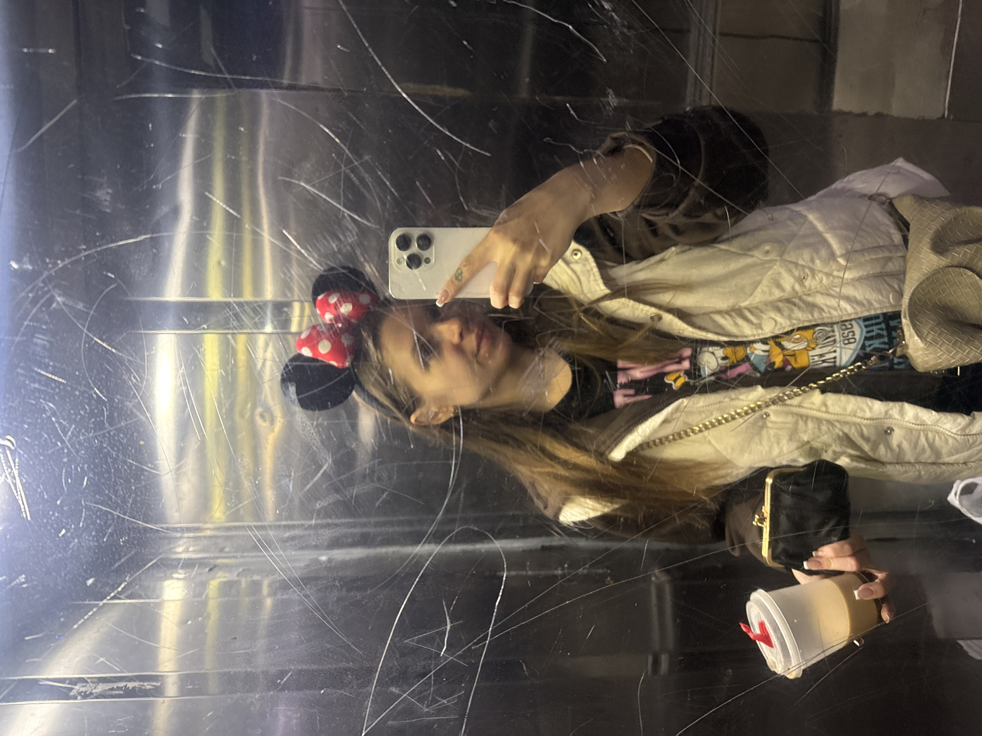 An indoor scene depicts a lone female adult taking a mirror selfie in what appears to be an elevator or a room with a highly reflective, heavily scratched metallic surface, likely located in Batumi, Georgia. The subject is a young to middle-aged woman with long brown hair, facing the mirror and holding a light-colored smartphone (possibly an iPhone with a triple-lens camera array) in her right hand to capture the image. She wears a black Minnie Mouse-style headband with a red and white polka-dotted bow. Her attire consists of a dark long-sleeved shirt with a multi-colored graphic design (featuring cartoon characters, possibly Disney), topped by a light beige or off-white quilted vest. In her left hand, she holds a clear plastic cup containing a light brown beverage, possibly coffee, with a red plastic stir stick or stopper protruding from the lid. Also in her left hand, positioned beneath the drink, is a small, black, framed coin purse or clutch. A light beige woven shoulder bag is visible hanging from her right shoulder, partially obscured. Her fingernails are manicured with French tips. The reflective surface she is photographing herself in exhibits numerous prominent vertical and diagonal scratch marks, along with minor smudges.