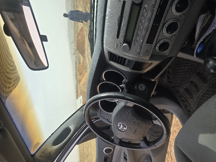 The image displays the interior of a vehicle, looking out through the windshield, with the scene identified as DU, Nigeria. The foreground shows the interior of a grey-colored Toyota vehicle, specifically the dashboard, steering wheel with a Toyota emblem, visible instrument cluster (two circular gauges), and a silver-toned center console containing a stereo unit and climate controls. Keys are present in the ignition, turned to an active position. A black, textured rubber floor mat is visible on the driver's side floor. A black leather-wrapped gear shifter for a manual transmission is in the lower right. The upper part of the windshield exhibits an amber tint, and a rearview mirror is positioned centrally.
Beyond the windshield, the environment appears arid and dusty under a hazy sky. Several conical piles of light brown earth or sand are present across the mid-ground, suggesting excavation or material handling. In the background, a single individual is observed walking away from the viewer, moving towards the right side of the frame. This person wears a loose-fitting, light-colored, long-sleeved garment. No other structures, vehicles, or significant vegetation are distinctly visible in the expansive outdoor area.