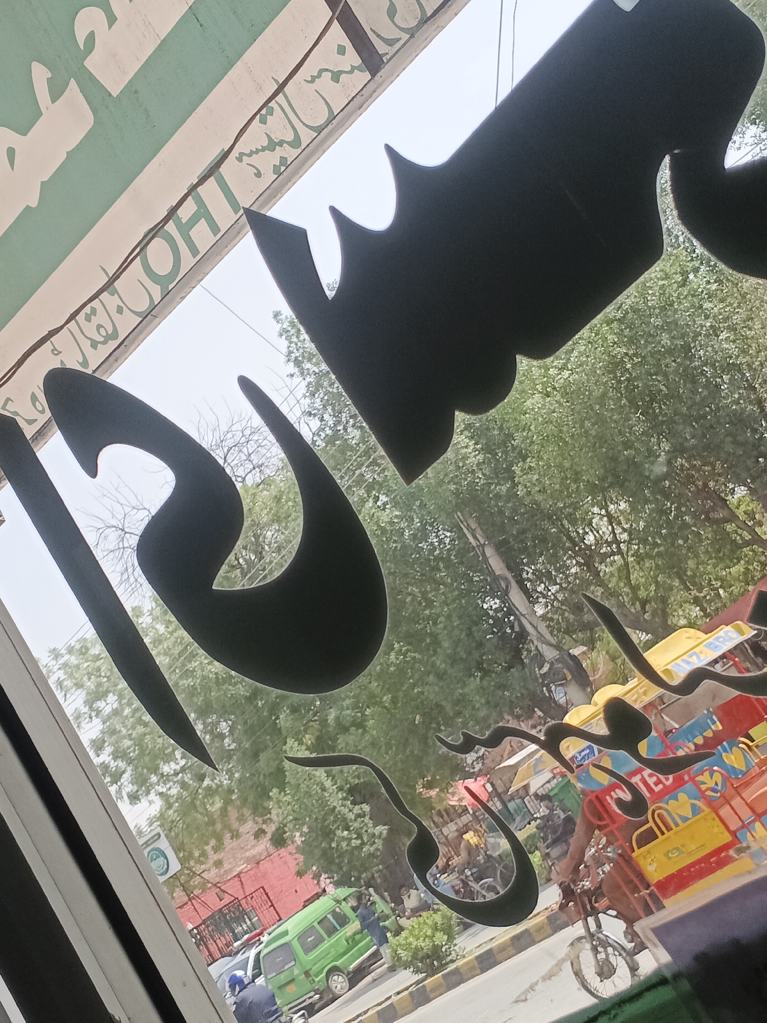 The image presents a street scene in Samundri, Pakistan, viewed from indoors through a window adorned with prominent, stylized calligraphic decals, likely in Arabic or Urdu script, cut from dark material. The camera is tilted, offering an angled perspective of the bustling outdoor environment.
Outside, the scene is vibrant and active under bright daylight. A street is visible with various vehicles and people. Notable vehicles include a bright green passenger van, a uniquely colorful auto-rickshaw-like vehicle (possibly a 'Chingchi' or 'tuk-tuk') painted in yellow, red, and blue, and a person on a motorcycle or bicycle. Text "HAZE BRO" and "UNITED" are visible on the side of the colorful vehicle, along with "17-380". People are seen on the street, near the vehicles, and going about their daily routines.
Lush green trees line the street, with some utility poles and overhead lines also present. In the midground, a building with a distinctive red brick wall is visible, along with another structure featuring a green sign that bears a circular white logo and some unreadable text. Above the window from which the photo is taken, a light green sign with white Arabic/Urdu script and partially visible English letters "OHT" can be seen, indicating a shop or establishment. The atmosphere suggests a typical busy day in a South Asian town.