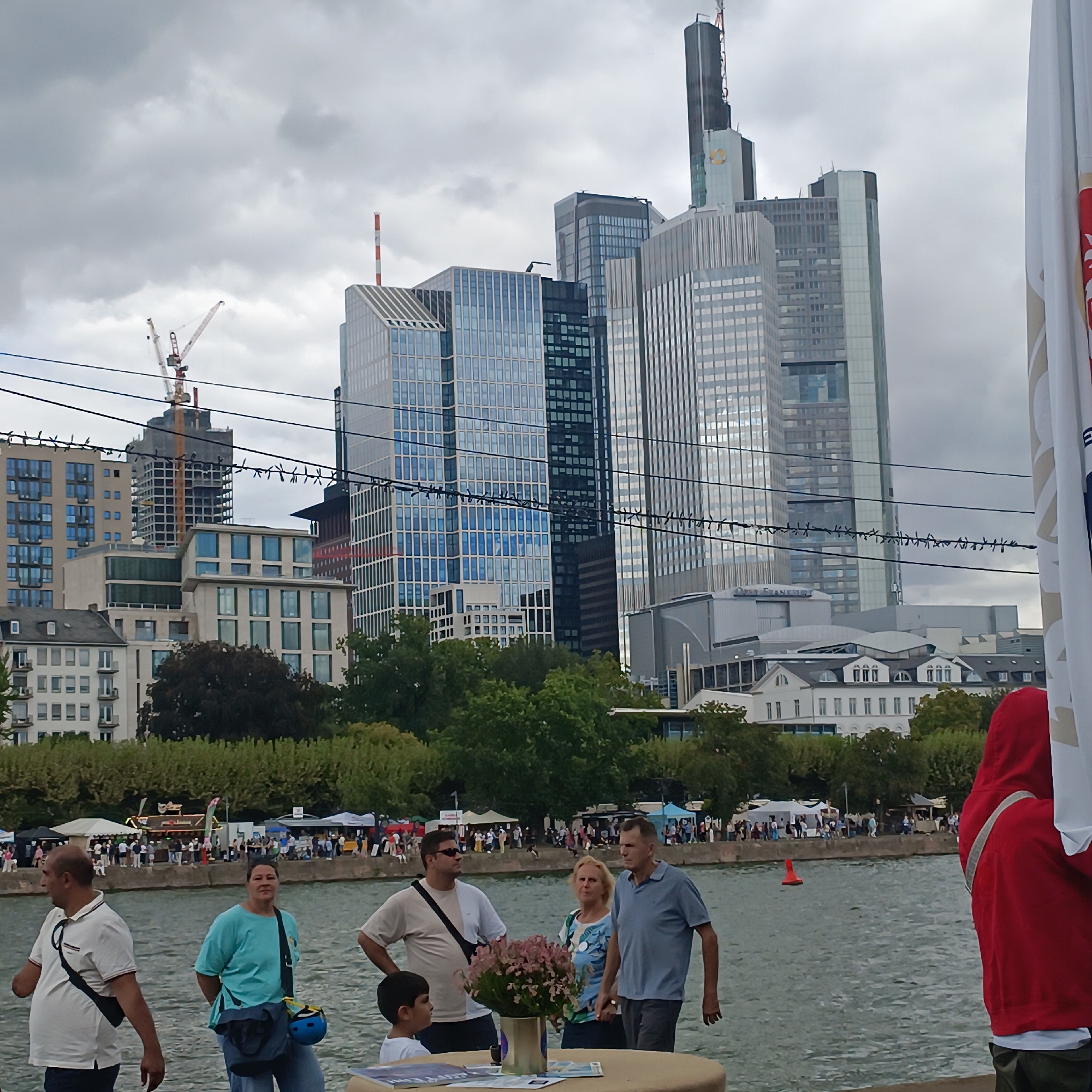 This is a medium shot of a waterfront scene in Frankfurt, Germany, with a prominent skyline of modern skyscrapers and a cloudy sky. In the foreground, a group of people are gathered near the water's edge. To the left, a man in a white polo shirt and dark pants is facing away from the camera, with a black strap across his chest. Next to him, a woman in a turquoise t-shirt and blue jeans is looking towards the camera. A young boy in a white shirt is to her left. Further to the right, a man in a blue polo shirt and dark pants is holding hands with a blonde woman in a patterned shirt. Behind them, a cluster of pink flowers in a vase sits on a table. In the background, across the water, there are stalls and tents set up along the embankment, suggesting an outdoor market or event. The river itself is calm and reflective. Dominating the background is the Frankfurt skyline, featuring several tall, glass-fronted buildings and a distinctive skyscraper with a dark spire topped by a red and white antenna. A construction crane is visible to the left of the main cluster of buildings. The overall atmosphere is that of a public gathering on a somewhat overcast day.