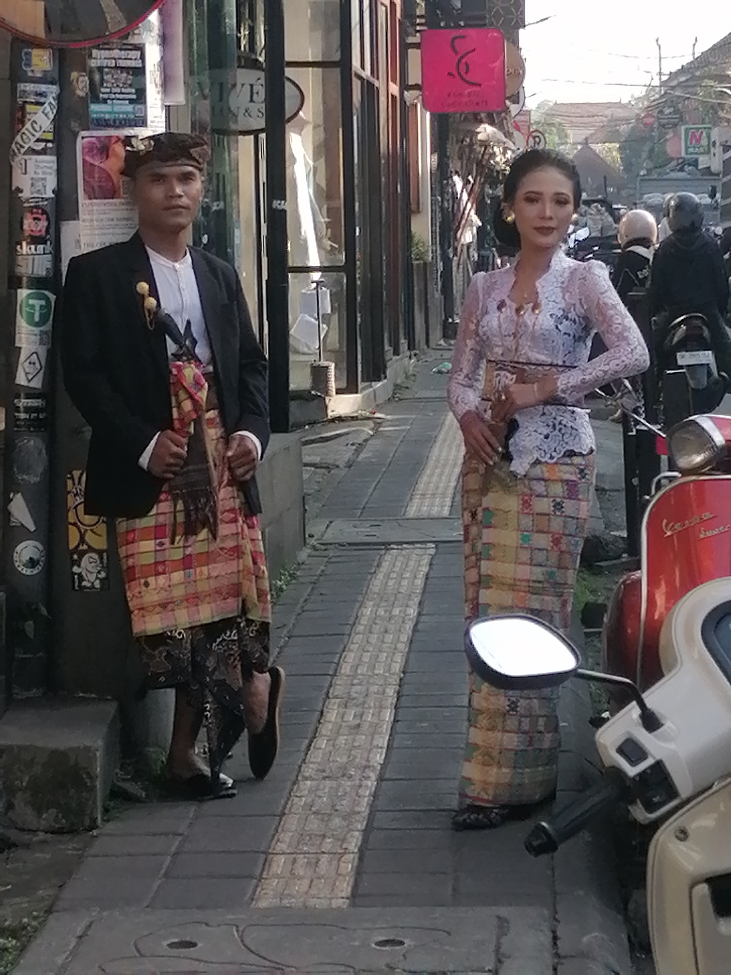 In Ubud District, Indonesia, a man and a woman stand on a paved sidewalk. The man, on the left, wears a black blazer over a white shirt and a patterned sarong, a traditional Balinese headdress, and dark shoes. He holds his hands clasped in front of him. The woman, on the right, wears a white lace top and a long, patterned skirt. She is adorned with jewelry and her hair is styled up. She stands with her hands clasped at her waist. A red scooter and a cream-colored scooter are parked to the right of the woman. The background shows buildings and other parked scooters. The scene appears to be outdoors, likely during the day.