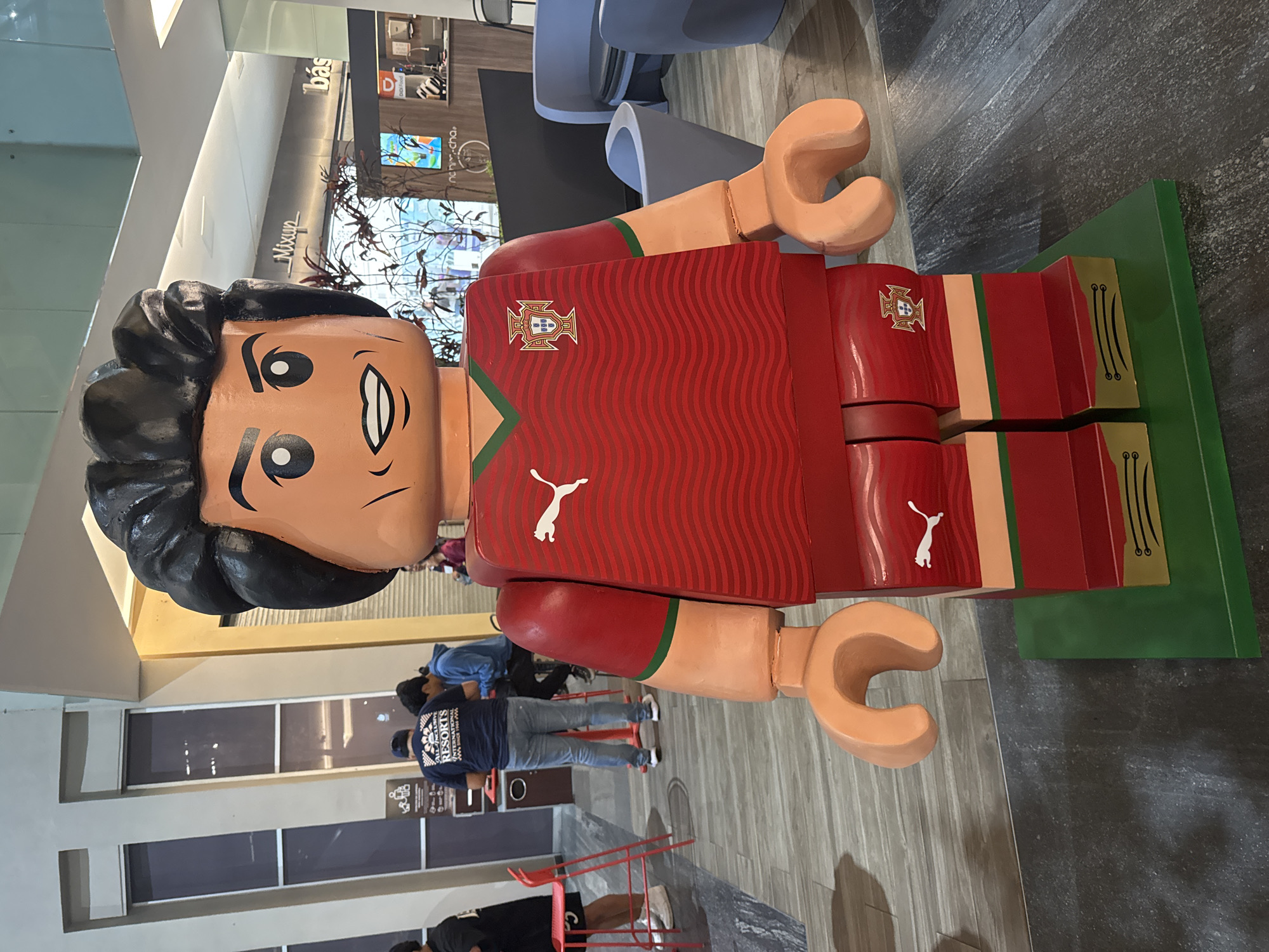 A full-body shot portrays a large LEGO figure of Cristiano Ronaldo standing in a shopping mall in Puebla, Mexico. The figure is wearing a red Portugal national team jersey with a white Puma logo. The jersey has a wave pattern and the Portuguese coat of arms emblem. The figure is standing on a green base. People are visible in the background. The mall's interior is visible in the shot.
