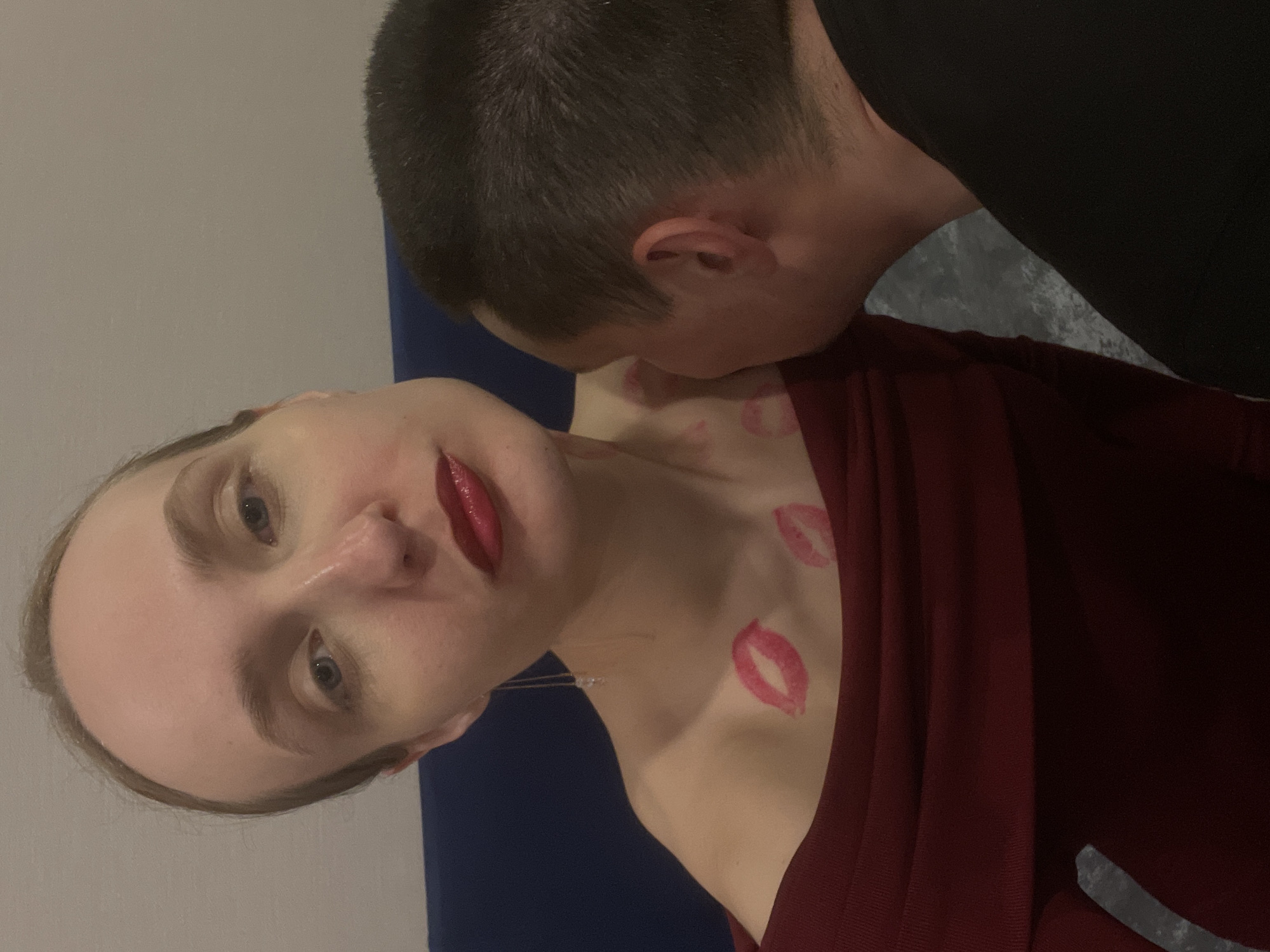 A man is kissing a woman on the neck. The woman is wearing a red dress and has red lipstick on. There are lipstick marks on her neck and chest. The background appears to be a room with a blue couch and gray wall. The location is stated to be Подшивалово, Russia.